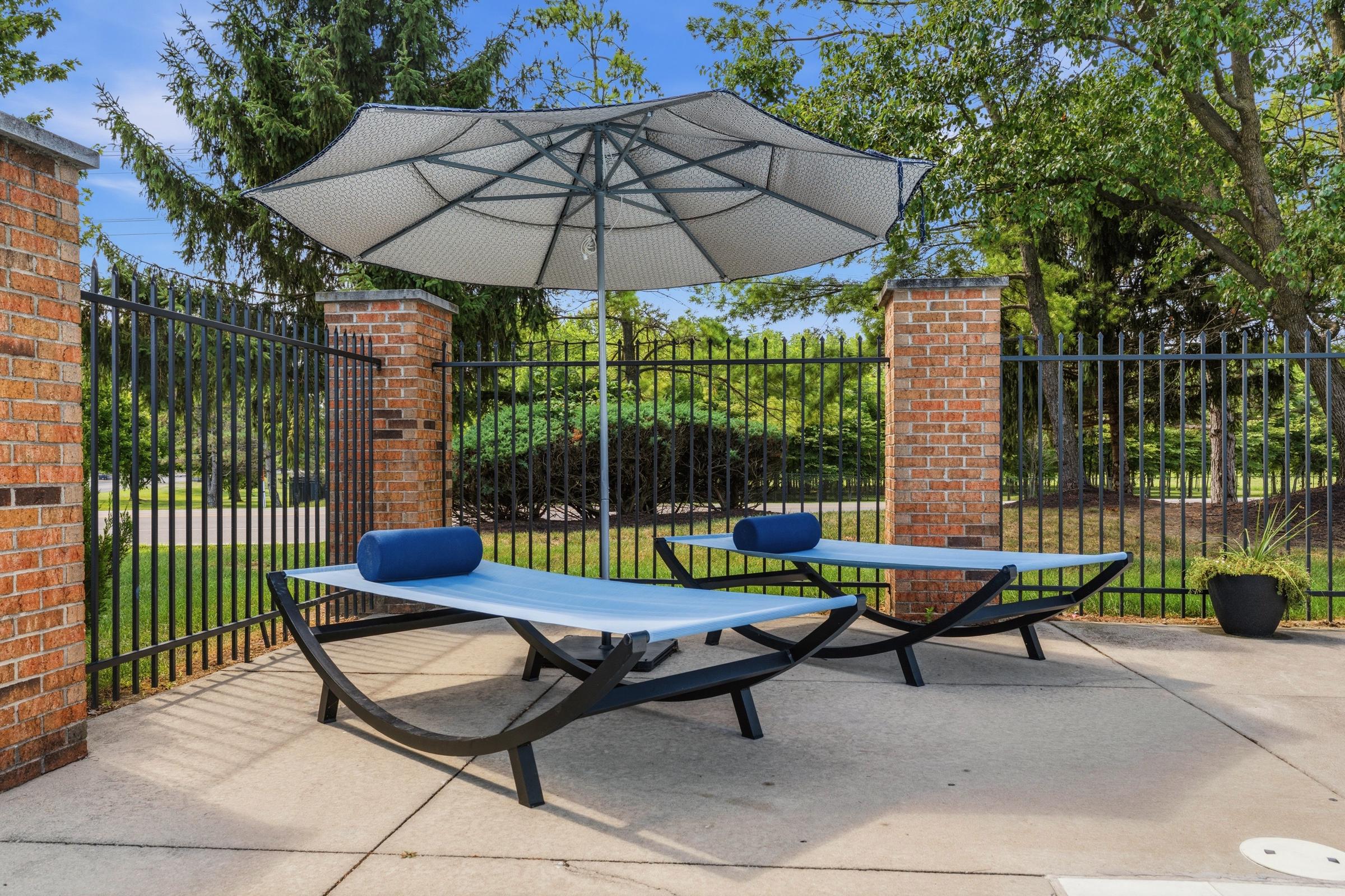 Two modern lounge chairs with blue cushions are positioned beneath a large gray umbrella. The setting is outdoors, featuring a paved patio area enclosed by black metal fencing and red brick pillars. Lush greenery and trees are visible in the background under a clear blue sky.