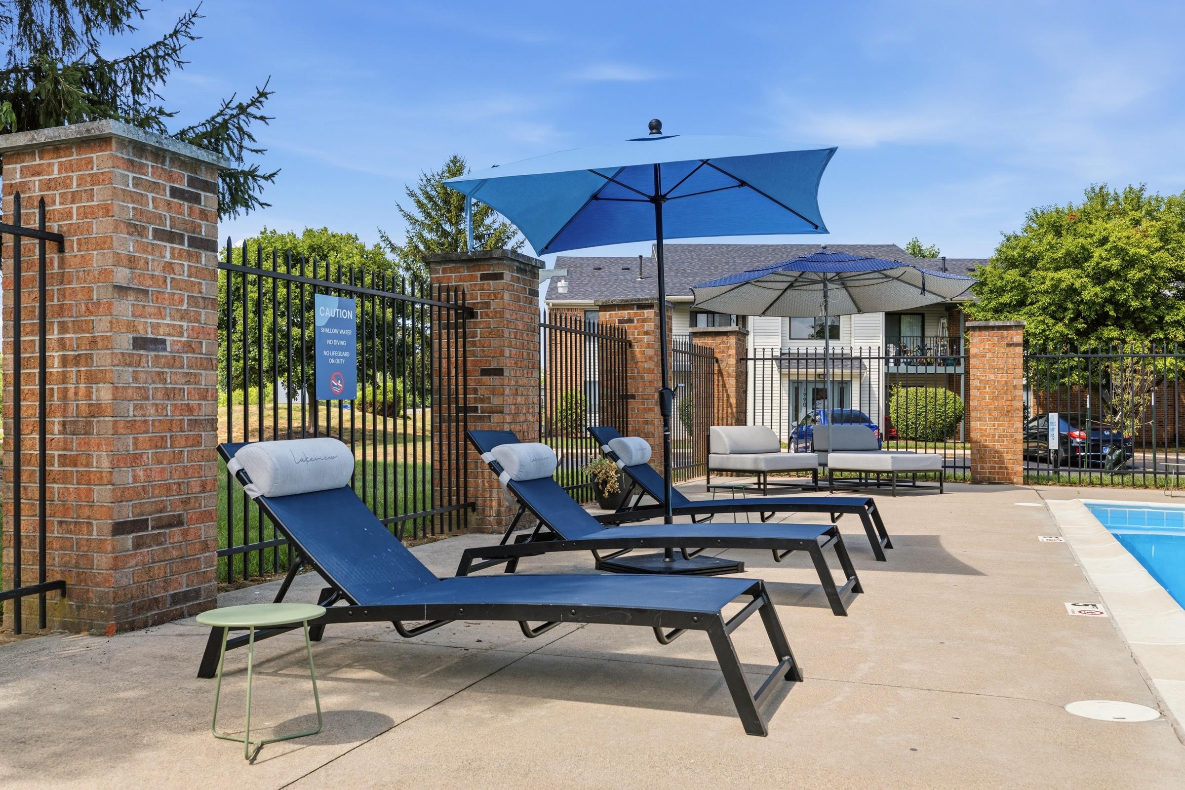 A sunny pool area featuring three lounge chairs with towels, an umbrella providing shade, and a small green side table. In the background, a gated fence and residential buildings are visible among trees and a blue sky. The atmosphere is relaxed and inviting for outdoor leisure.