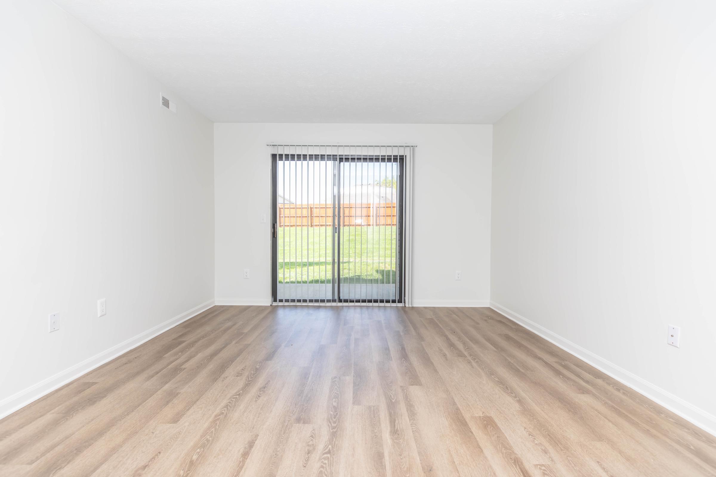 Empty room with light-colored walls and wooden flooring. A sliding glass door with vertical blinds provides access to an outside area, with a view of a grassy lawn and fence. The room has no furniture, creating a spacious and airy feel.