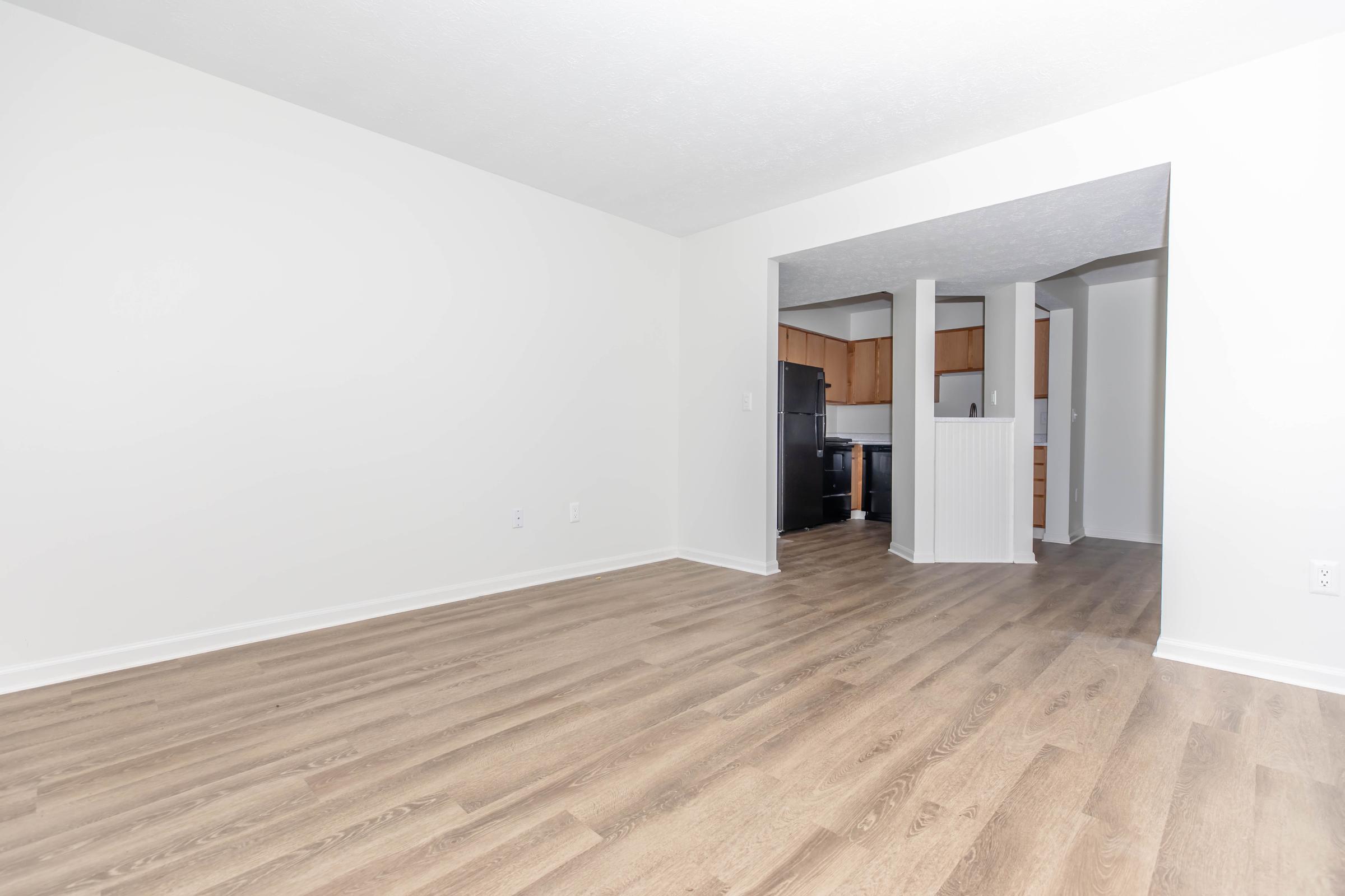 Empty room featuring light-colored walls and a wood laminate floor. An open archway leads to a kitchen with wooden cabinets and black appliances. The space is well-lit and unadorned, creating a blank canvas for potential decoration.