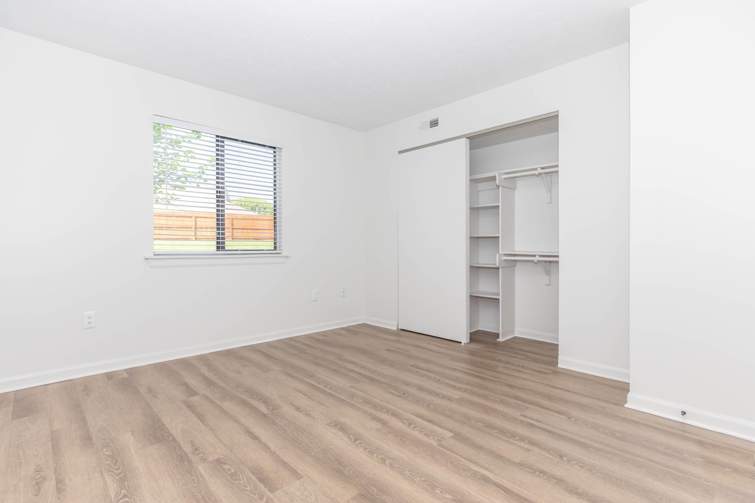 A bright, empty room with white walls, wood flooring, and a window featuring horizontal blinds. A closet with sliding doors is visible on one side, and the room has no furniture, creating a spacious and clean atmosphere. Natural light filters in through the window, enhancing the overall brightness.