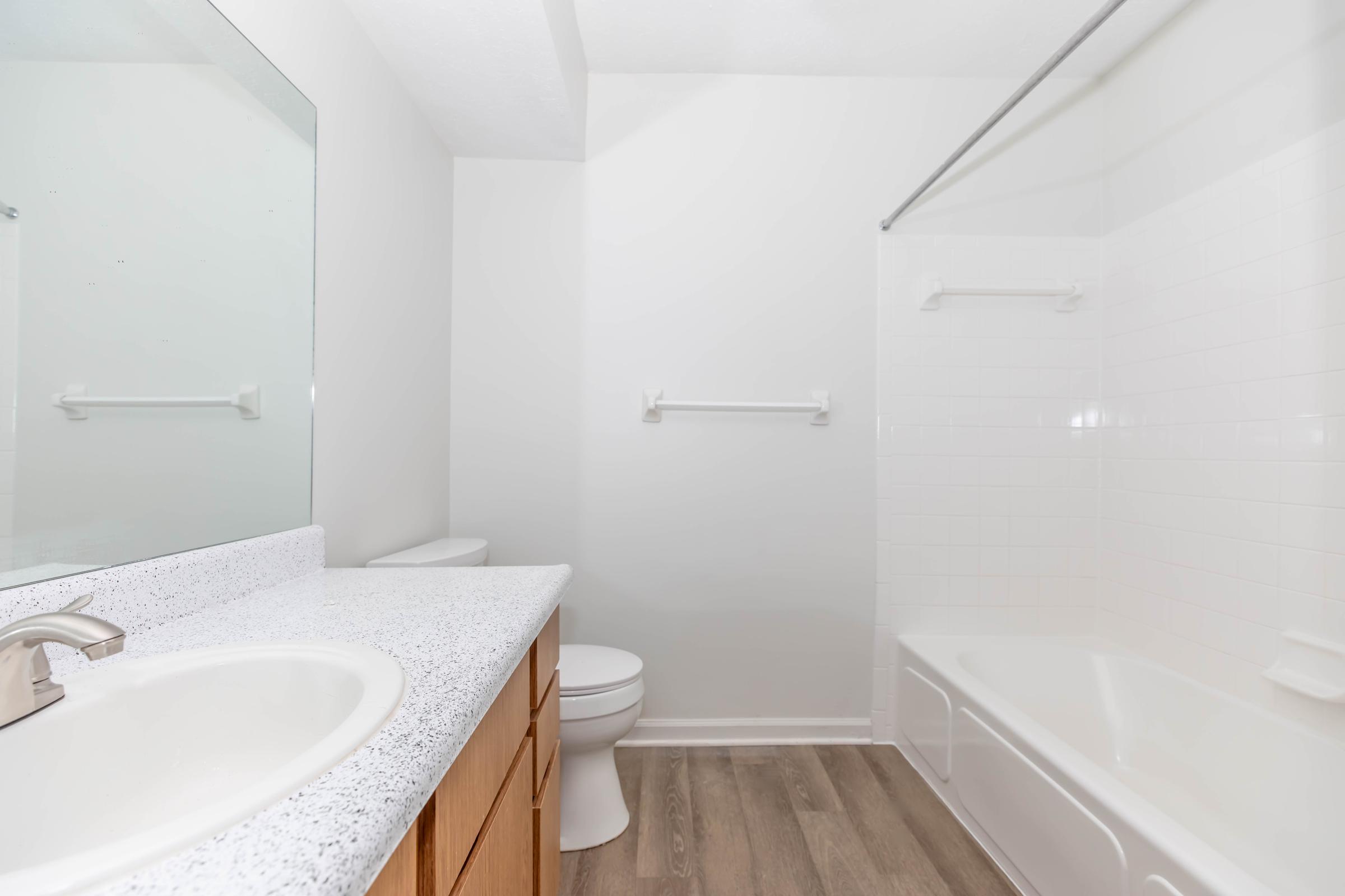 A clean and bright bathroom featuring a white bathtub with a shower, a large mirror above a light-colored countertop with a sink, and a toilet. The walls are painted in a light color, and there is a towel rack. The flooring is a light brown wood-like material, creating a modern and spacious appearance.