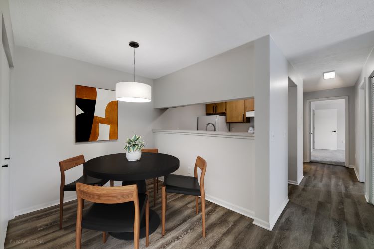 A modern dining area featuring a round black table with four wooden chairs. A pendant light hangs above the table. A colorful abstract painting is on the wall, and a small plant adds a touch of greenery. The room has muted walls and a view of a kitchen area in the background.