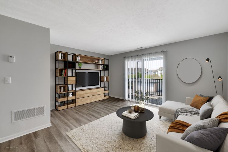 A modern living room featuring a light-colored sofa with orange and gray throw pillows, a circular black coffee table, and an entertainment unit with shelves. The room includes large windows with vertical blinds, allowing natural light, and has neutral gray walls and wooden flooring.