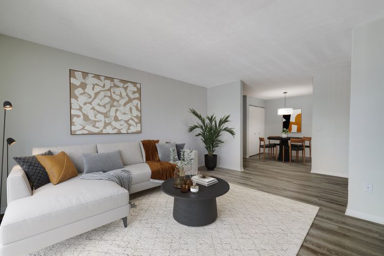 A modern living room featuring a light gray sectional sofa adorned with orange and gray cushions. A round black coffee table sits on a textured area rug, with decorative items on top. A plant stands nearby, and a large abstract artwork hangs on the wall. In the background, a dining area with a round table and chairs is visible.