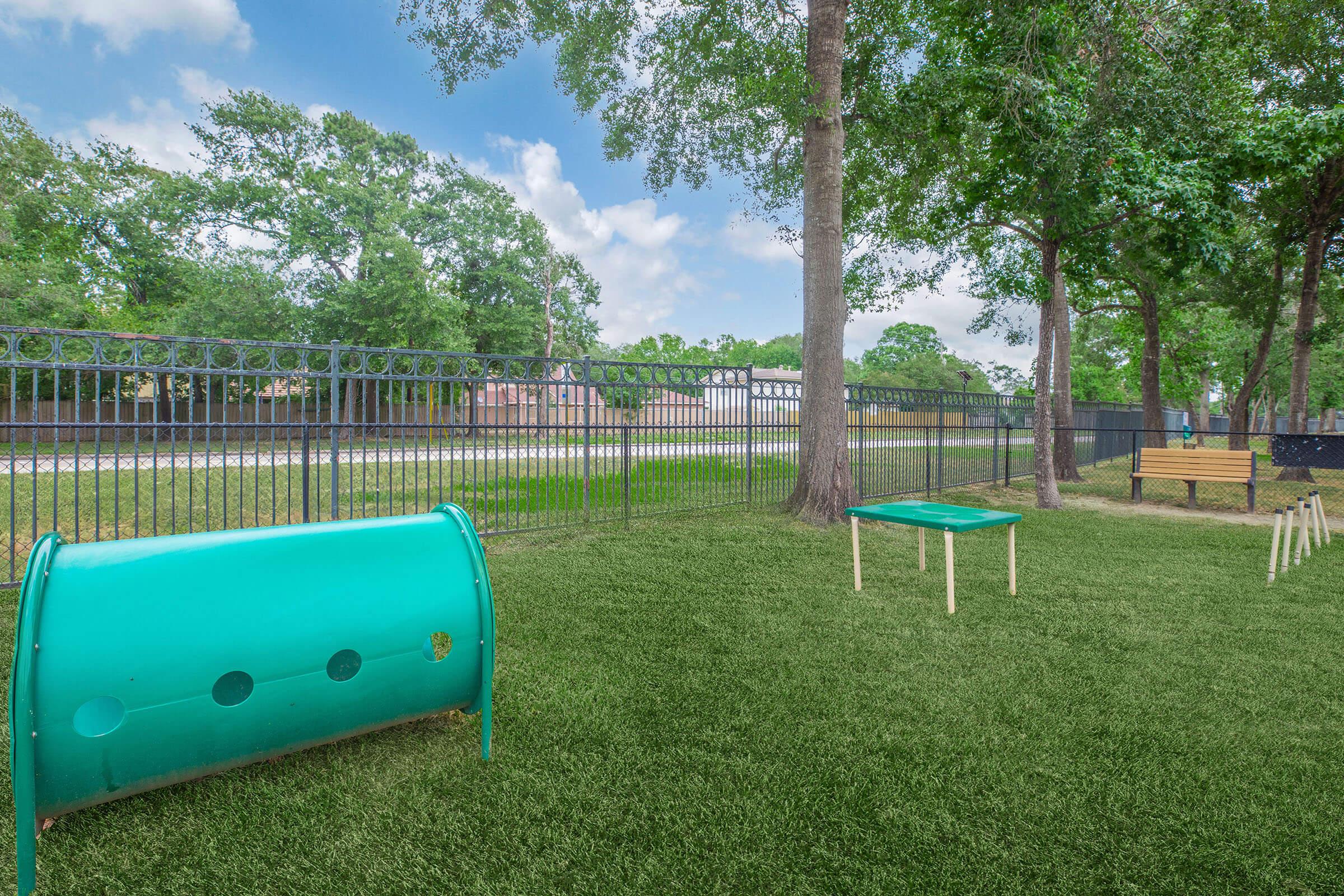 A green play area featuring a tunnel and a table on artificial grass, surrounded by trees and a black fence. In the background, there’s a view of a road and houses. The setting appears inviting and suitable for outdoor activities.