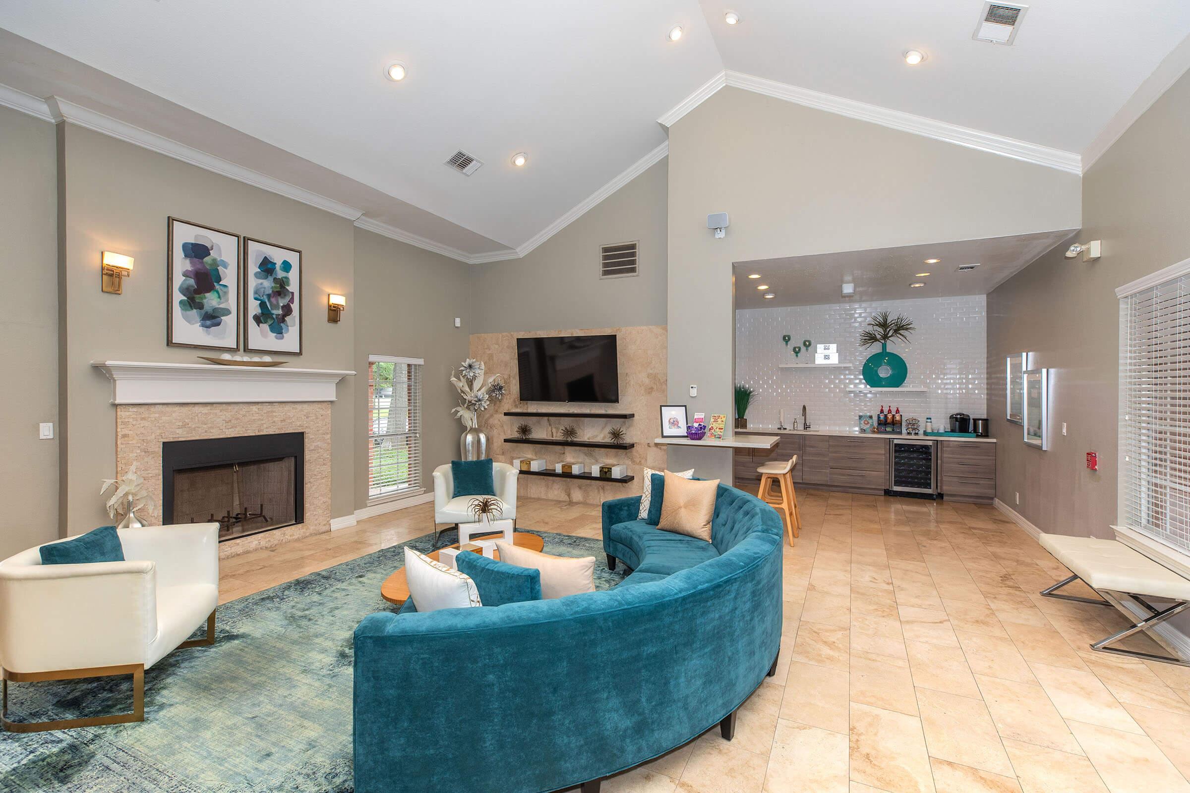 A modern lounge area featuring a cozy seating arrangement with teal and white chairs, a fireplace, and a wall-mounted TV. A stylish bar area with a counter, decorative elements, and natural light filters in through large windows, creating an inviting atmosphere.