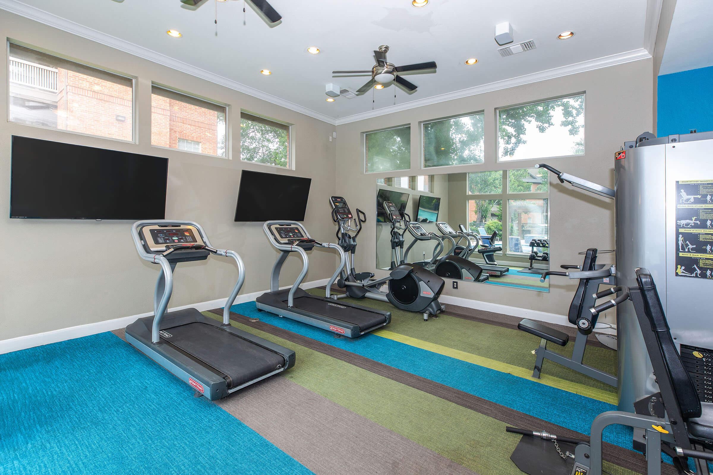 A modern fitness room featuring two treadmills, a stationary bike, and various exercise equipment. The space is well-lit with natural light from large windows and has a colorful, patterned carpet. Two flat-screen TVs are mounted on the wall for entertainment during workouts.