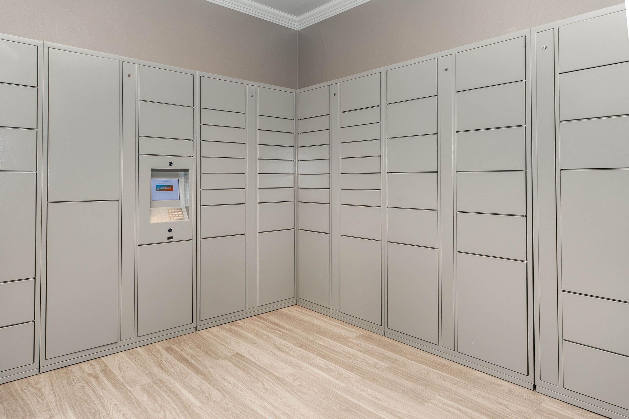 Interior view of a room featuring a wall of gray parcel lockers with a digital keypad and screen. The lockers are neatly arranged in rows, and the floor is made of light-colored wood. The walls are painted in a soft, neutral color, creating a modern and organized space for package delivery.