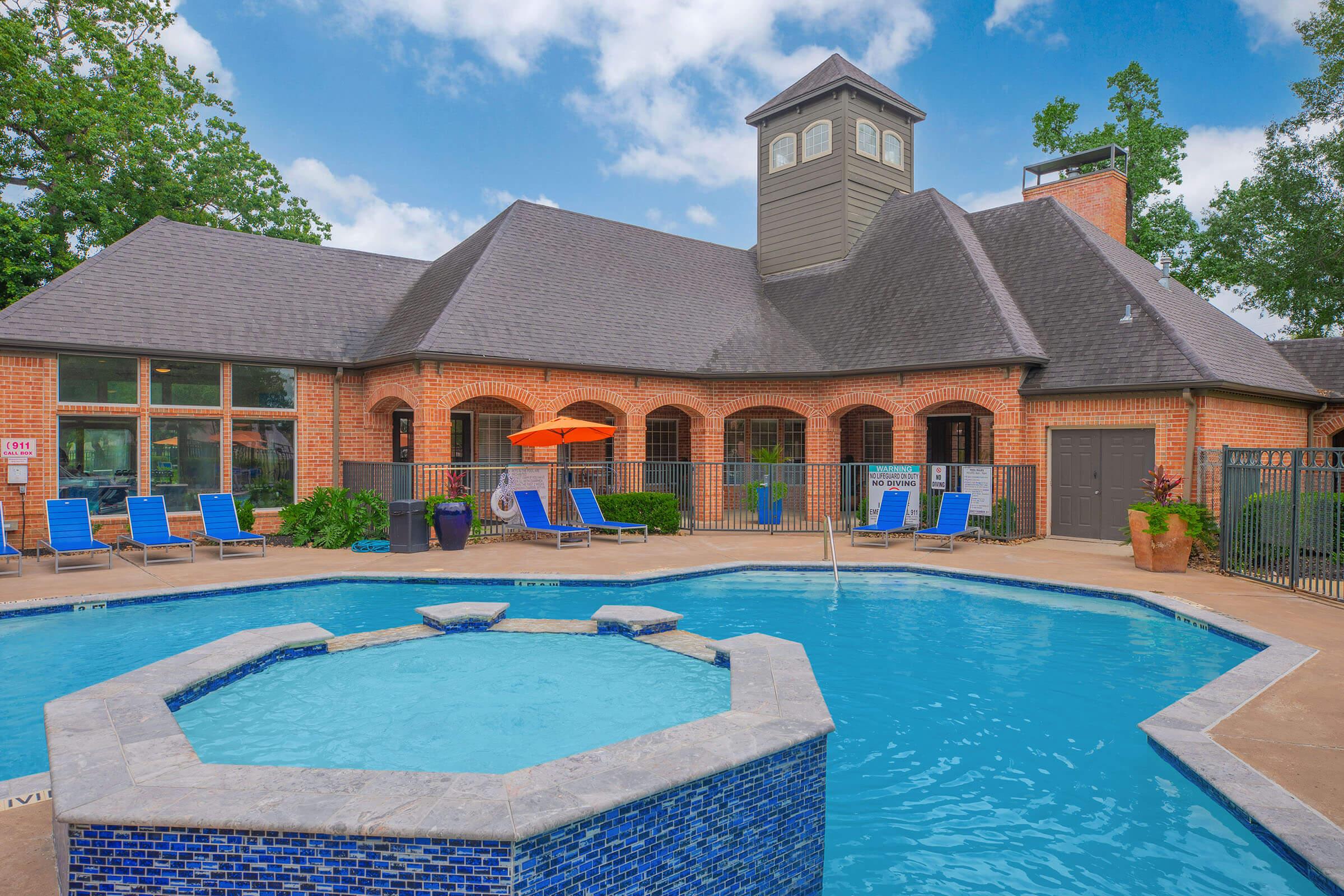 A well-maintained swimming pool surrounded by lounge chairs, featuring a hot tub in the foreground. In the background, there is a large brick building with a peaked roof and large windows, shaded by trees. An orange umbrella adds a pop of color to the outdoor area. Bright blue sky enhances the inviting atmosphere.
