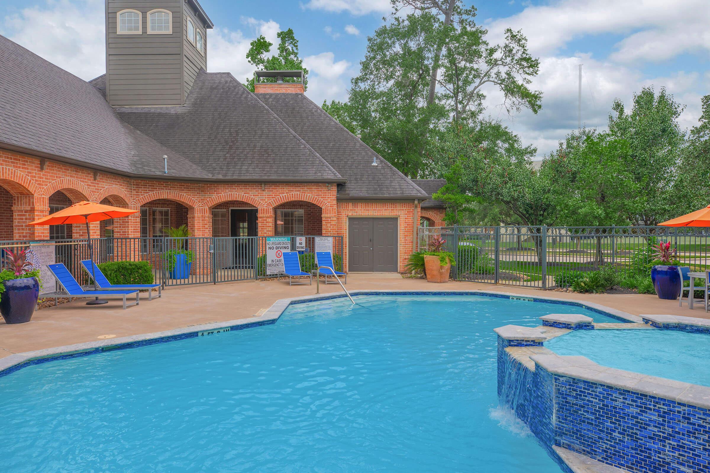 A tranquil outdoor swimming pool surrounded by lounge chairs and vibrant orange umbrellas. The pool features a stone waterfall and is located near a brick building with large windows. Lush green trees and a well-maintained lawn are visible in the background under a partly cloudy sky.
