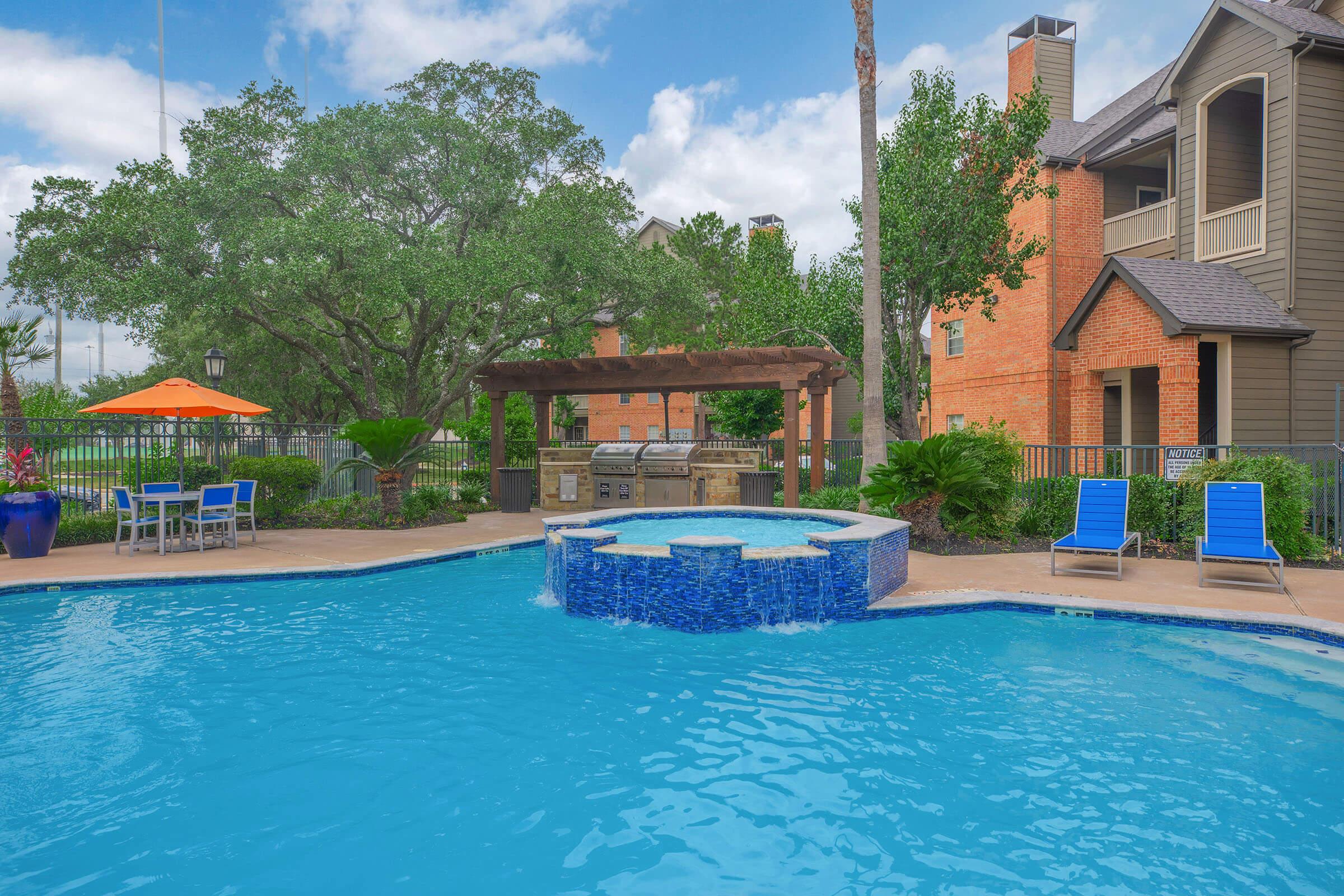 A bright outdoor pool area featuring a clear blue pool with a spa section, surrounded by lush greenery. There is a barbecue grill area under a pergola, along with several blue lounge chairs and an umbrella. In the background, there are apartment buildings and palm trees under a partly cloudy sky.