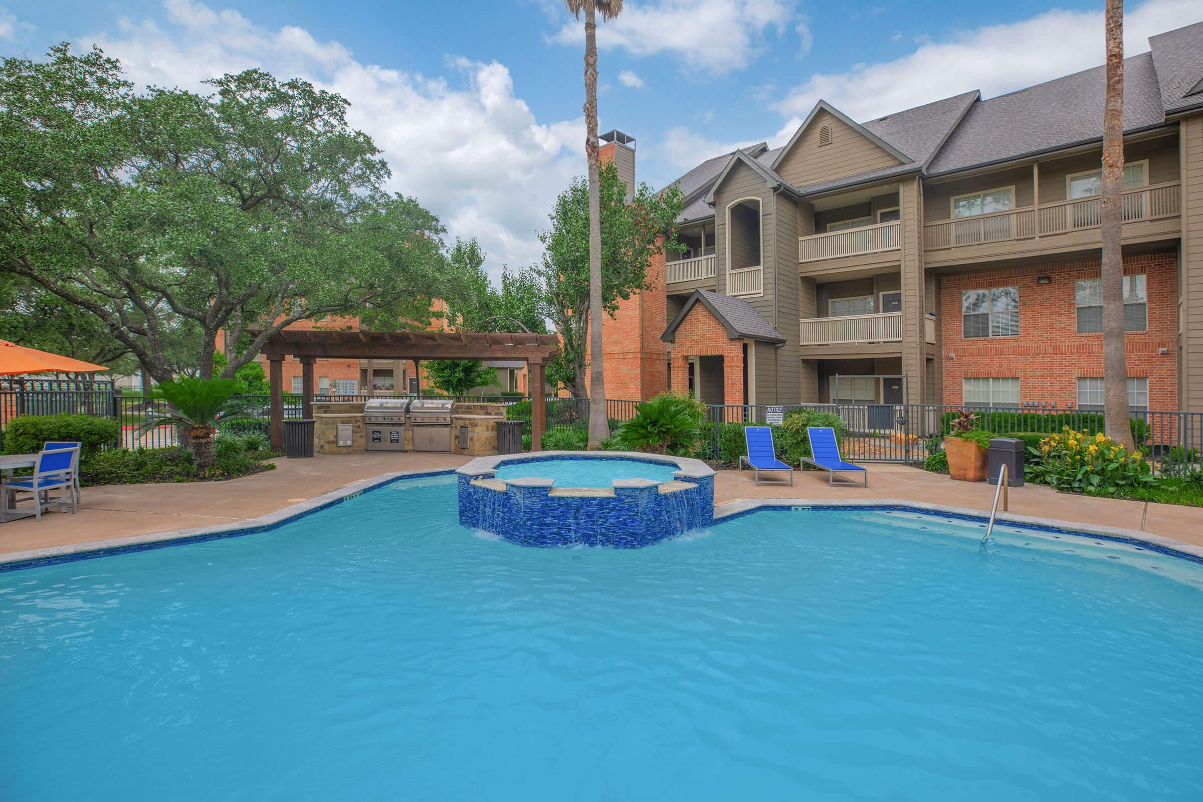 A vibrant pool area with a circular hot tub at the center, surrounded by lush greenery and palm trees. Comfortable lounge chairs are set around the pool, and a barbecue area is visible nearby. In the background, a multi-story apartment building is partially seen under a clear blue sky.