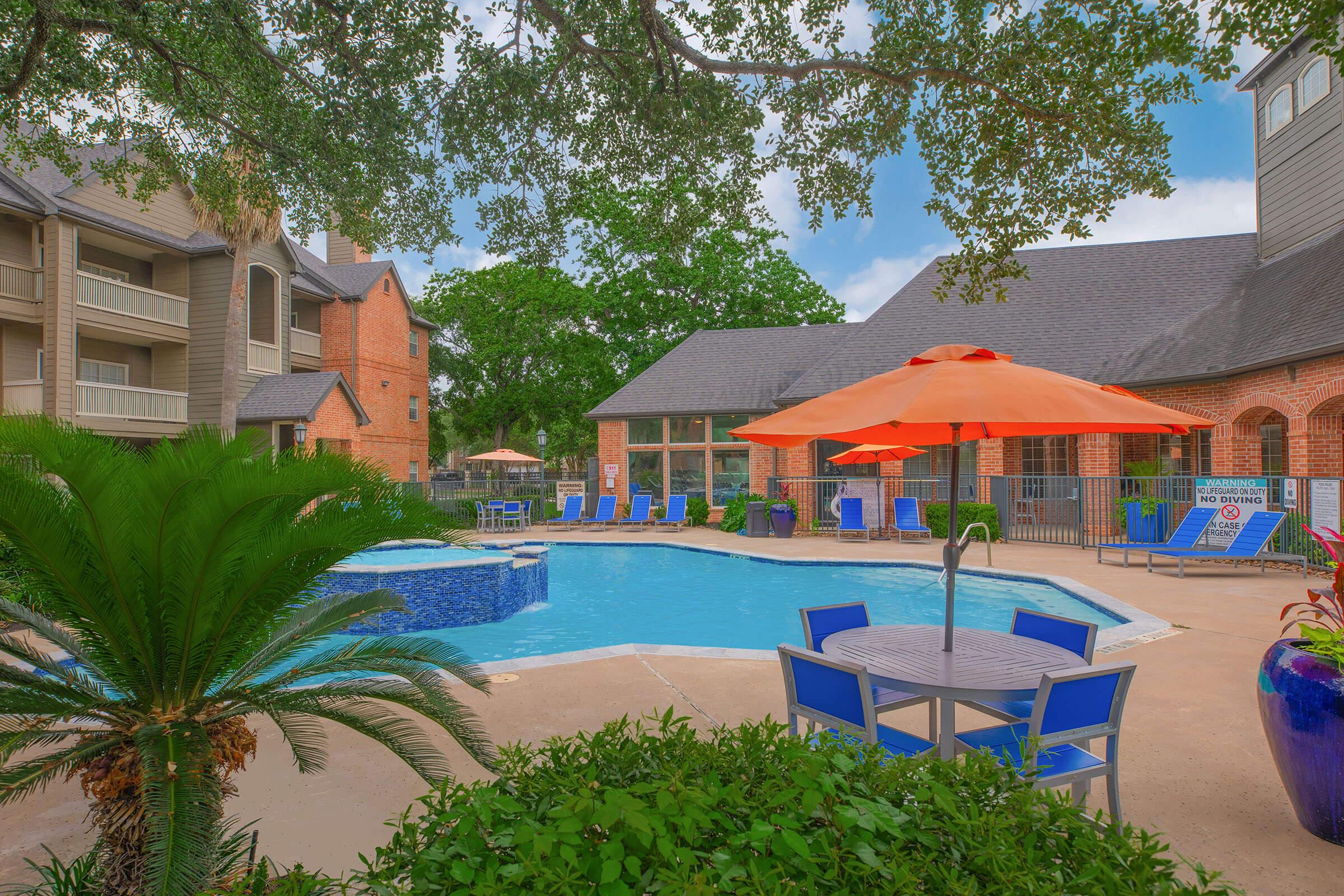 A bright and inviting swimming pool surrounded by lush greenery, orange umbrellas, and blue lounge chairs. In the background, there are buildings with a mix of brick and modern architecture. The scene is tranquil and designed for relaxation, with clear blue skies overhead.