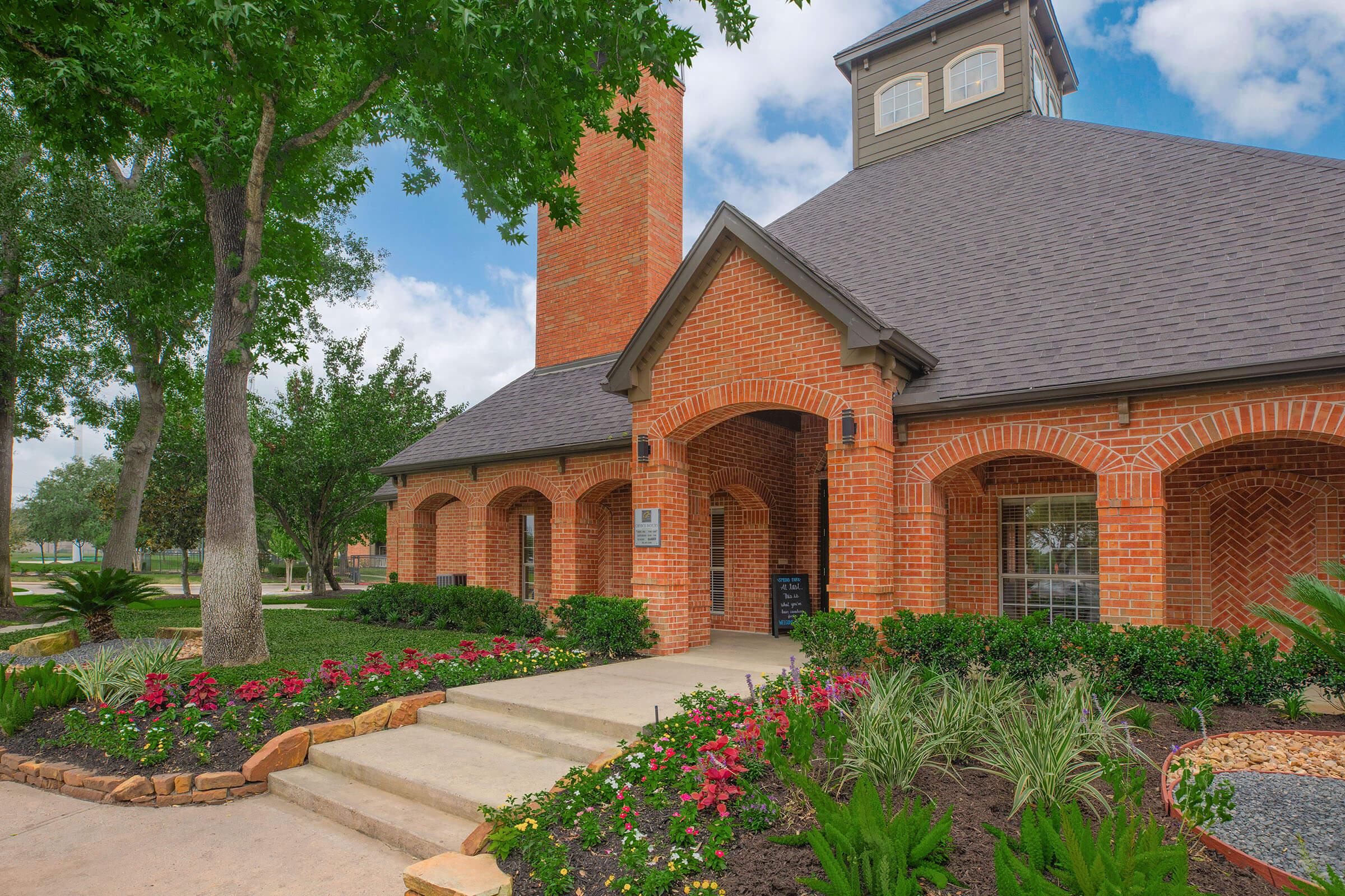A charming brick building with a pitched roof and a tall chimney, surrounded by lush green landscaping. Colorful flowerbeds line the walkway leading to the entrance, framed by trees and shrubs. The sky is partly cloudy, adding to the inviting atmosphere of the property.