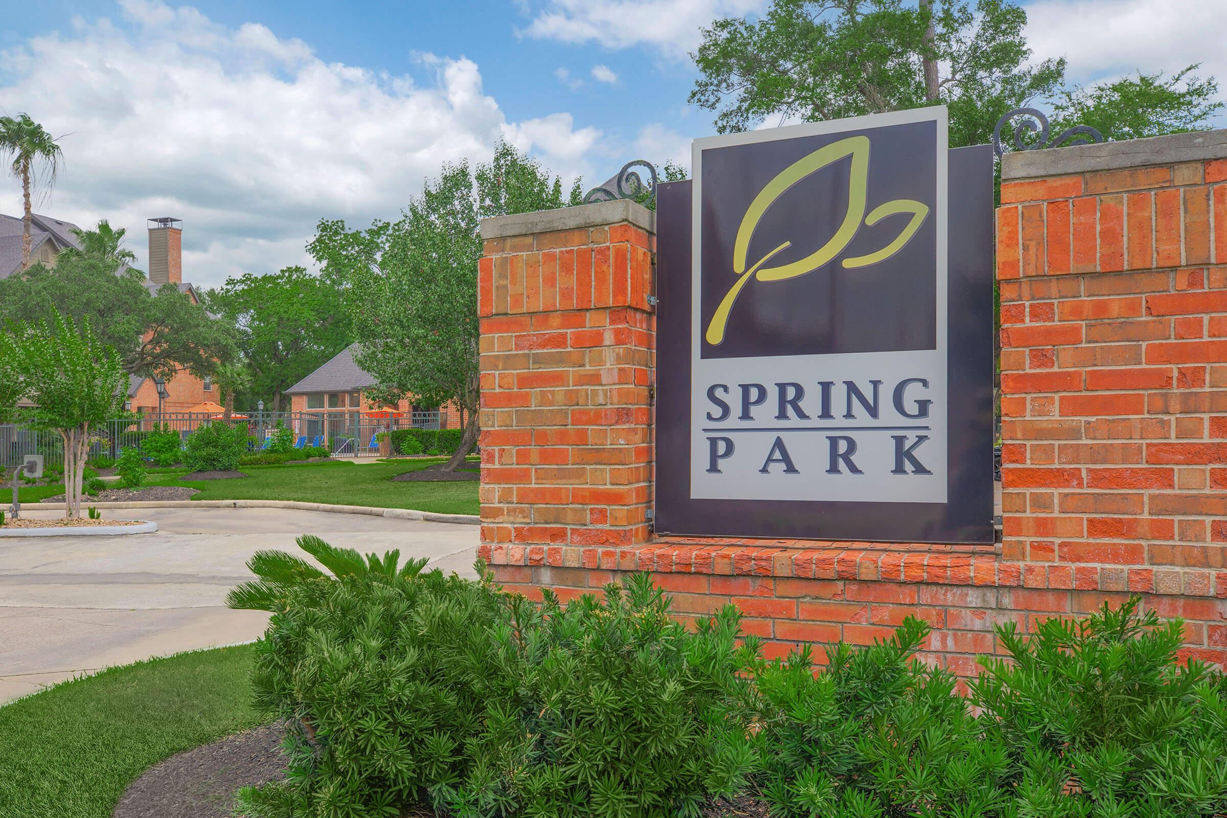 Sign for Spring Park featuring a green leaf logo, surrounded by landscaped greenery. The area has a well-maintained pathway and residential buildings visible in the background under a partly cloudy sky.