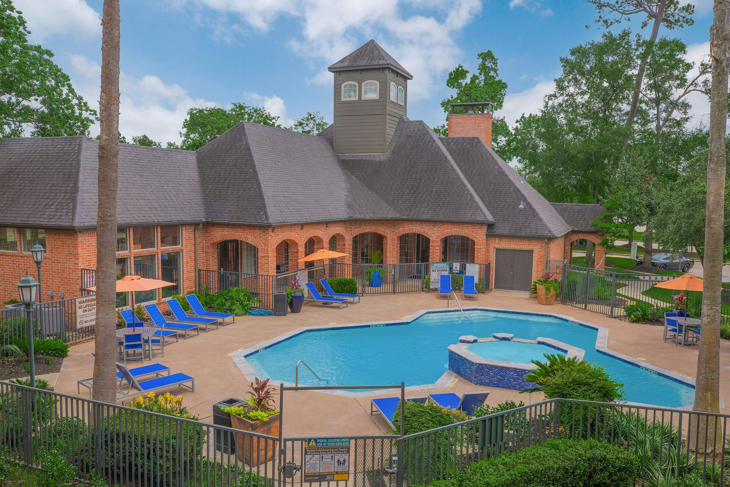 Aerial view of a resort-style pool area featuring a large blue swimming pool, a hot tub, lounge chairs, and umbrellas. Surrounding the pool are well-maintained shrubs and trees, with a clubhouse in the background showcasing a modern architectural design. Clear skies and a lush landscape enhance the inviting atmosphere.