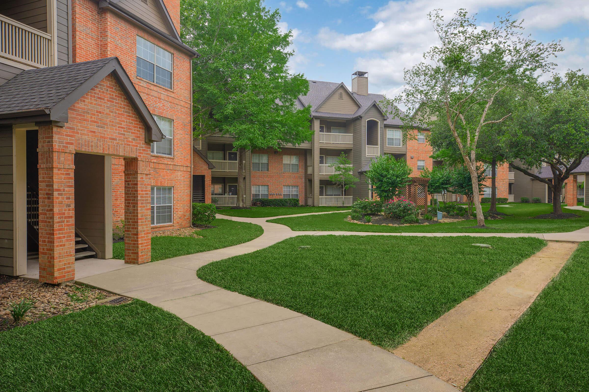 Lush green lawn with a well-maintained pathway winding through an apartment complex. Red brick buildings with balconies are visible on either side, surrounded by trees and shrubs. The sky is partly cloudy, creating a serene and inviting atmosphere.