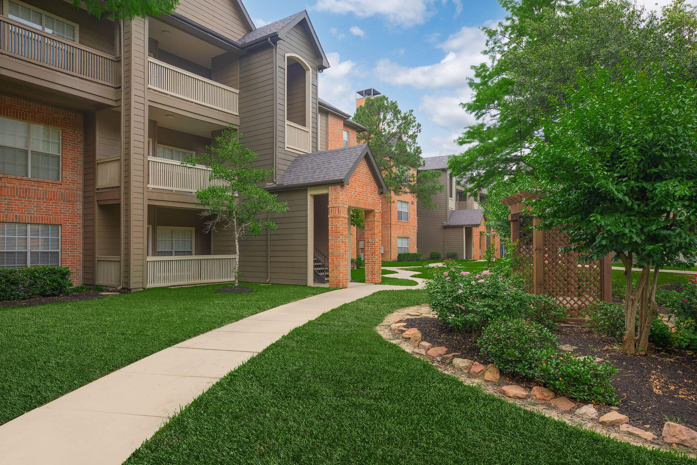 A well-maintained apartment complex with three buildings, surrounded by lush green grass and neatly trimmed shrubs. The pathway is paved, leading through the landscaped area with a stone border, creating an inviting atmosphere in a residential setting. The sky is partly cloudy, enhancing the peaceful environment.