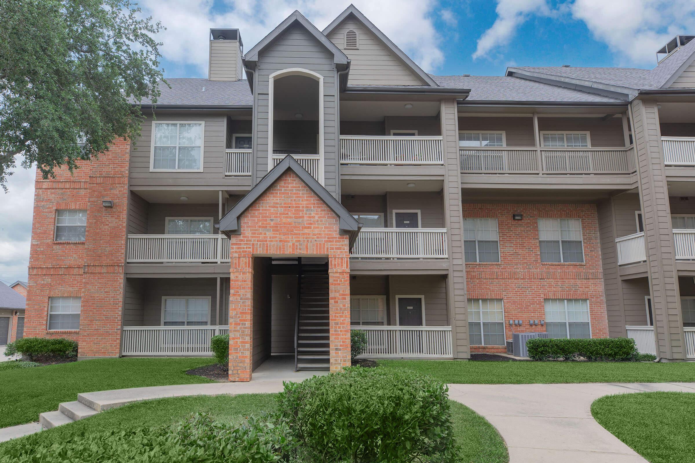 A multi-story residential building featuring brick and siding exteriors. The entrance has a covered archway leading to a staircase. Balconies are visible on the front and sides of the building, surrounded by well-maintained green grass and landscaping under a partly cloudy sky.