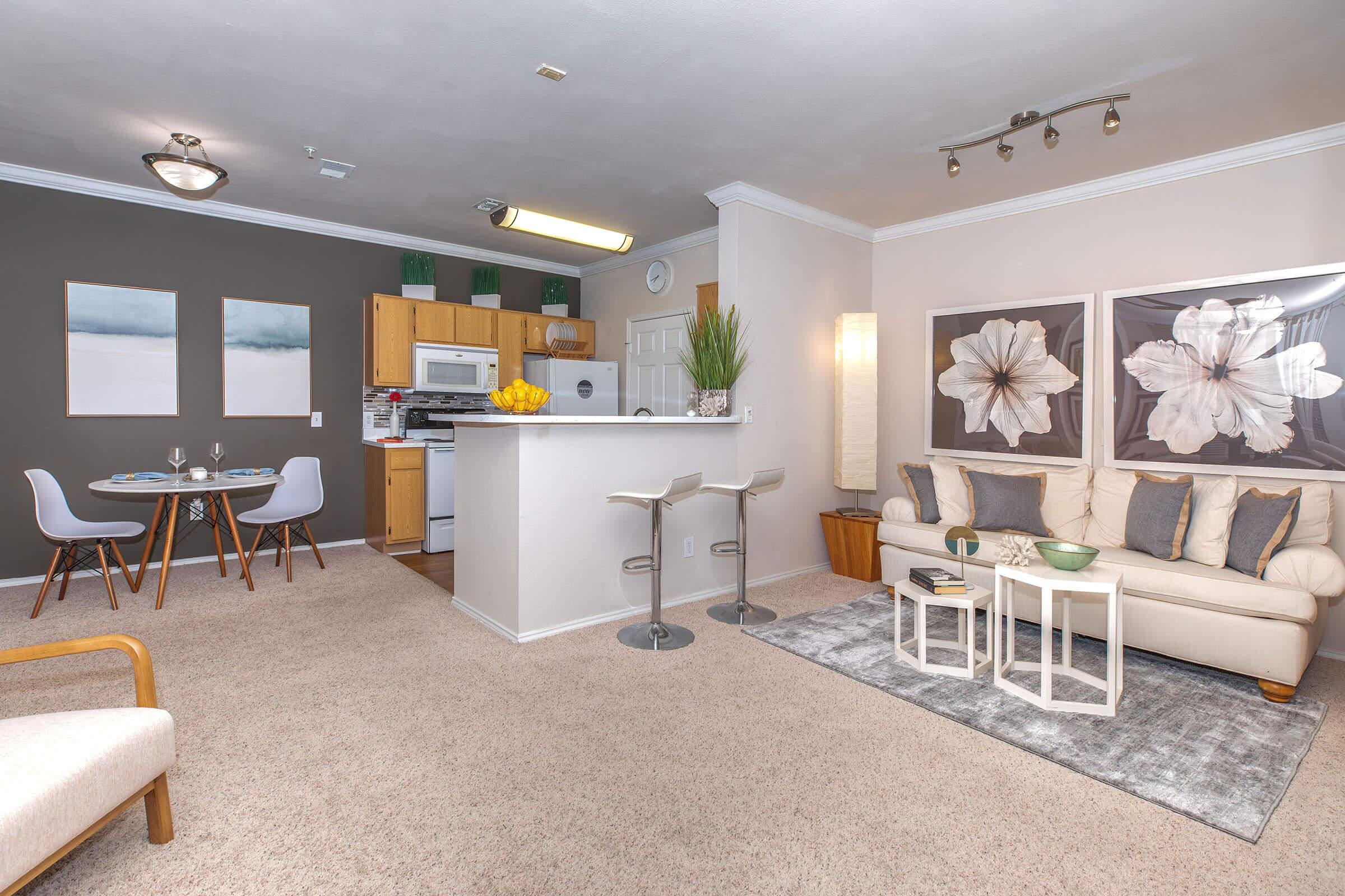 A modern, cozy apartment with an open layout. The living area features a beige sofa, decorative tables, and a rug, while the dining area includes a small table and chairs. The kitchen is equipped with wooden cabinets and white appliances. Two floral artworks adorn the walls, contributing to the stylish decor.