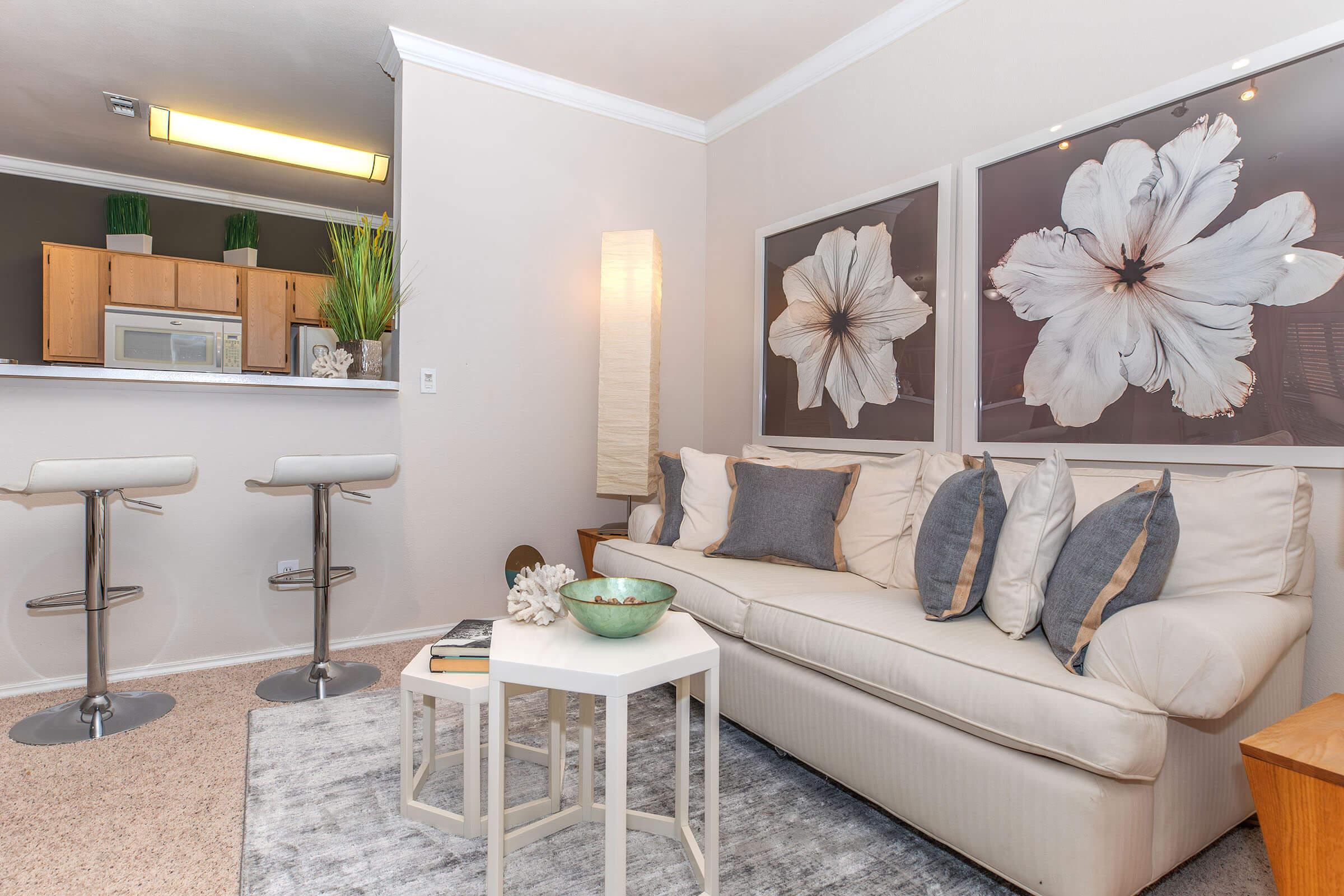 A bright and modern living room featuring a white sofa with decorative pillows, a small white coffee table, and a rug on the carpeted floor. Two bar stools are positioned at a kitchen counter in the background. Large floral artwork hangs on the walls, with greenery in the corner adding a fresh touch.