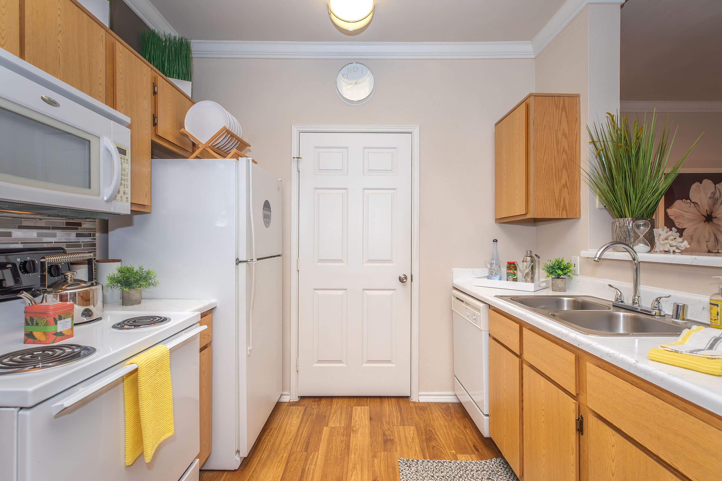 A clean and modern kitchen featuring wooden cabinets, white appliances including a microwave, refrigerator, and dishwasher. The countertop is equipped with a sink, and there are small plants and decorative items. A clock is mounted on the wall, and a door leads to another room. The floor is made of warm-colored wood.