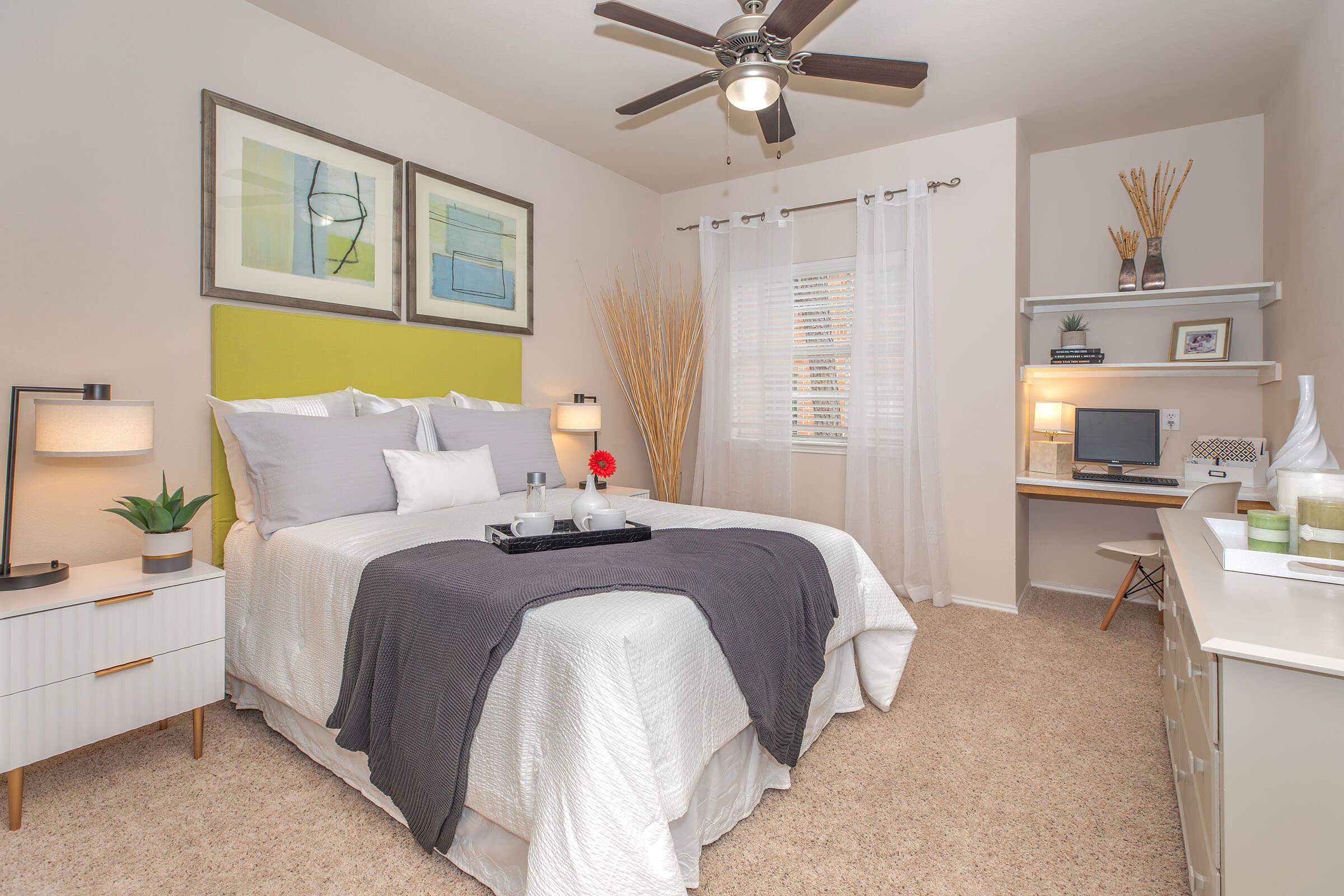 Cozy bedroom featuring a queen-sized bed with gray and white bedding, decorative pillows, and a bright green headboard. A ceiling fan and two lamps provide lighting, while a small desk and chair, along with decorative plants and framed artwork, enhance the inviting atmosphere. Natural light filters through sheer curtains.