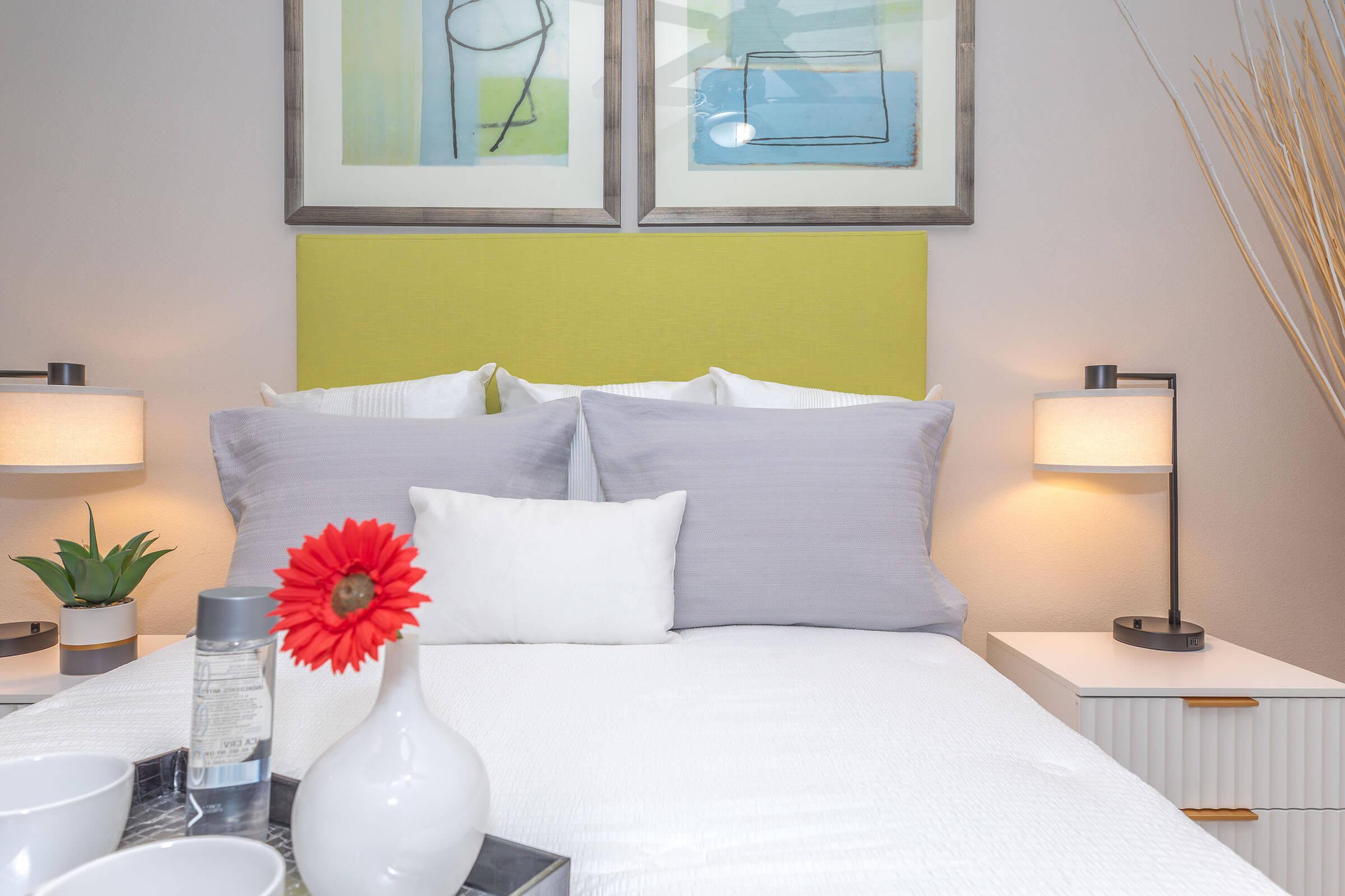 A cozy bedroom featuring a bed with gray and white pillows, a bright green headboard, and two bedside lamps. A small plant, a vase with a red flower, and a tray with a bottle and cups are arranged on a nightstand beside the bed. Abstract artwork adorns the wall above the headboard.
