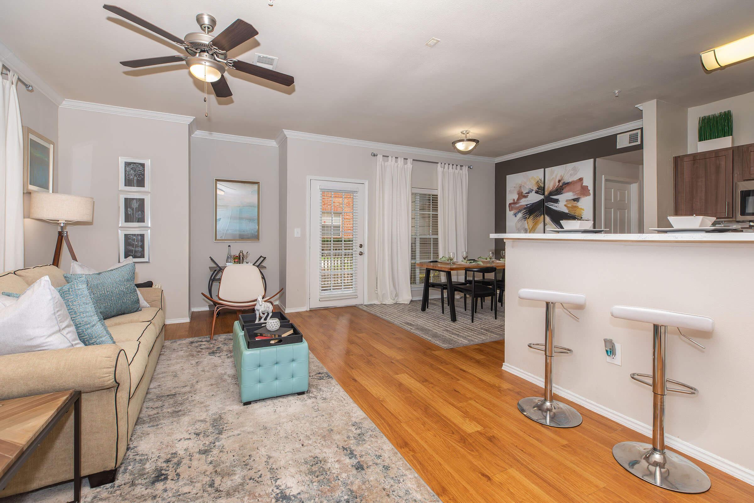 A cozy living area featuring a beige sofa with decorative pillows, a turquoise coffee table, and a stylish rug. There’s a ceiling fan above, a dining area with a wooden table, and modern bar stools. Large windows allow natural light, and artwork adorns the walls, creating a welcoming atmosphere.