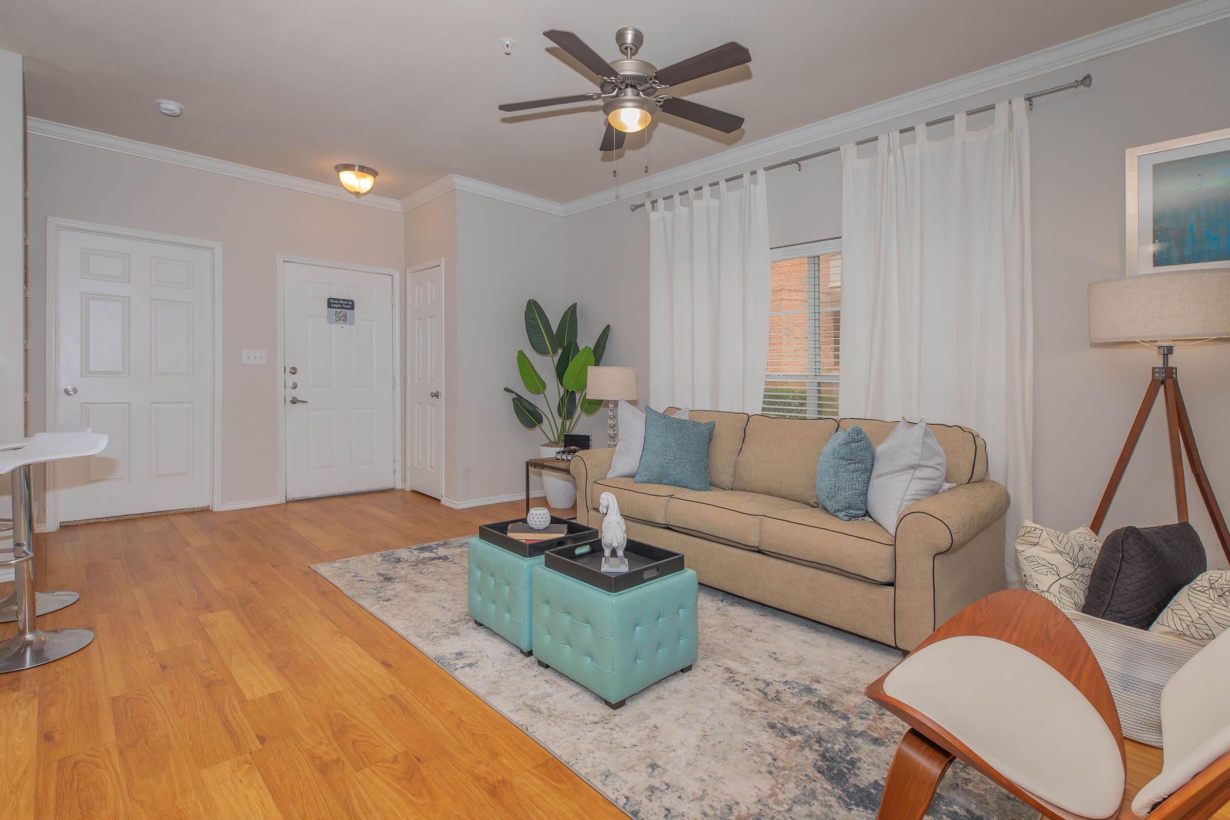 A cozy living room featuring a beige sofa with colorful cushions, a light blue coffee table, and a decorative area rug. There is a lamp and a plant beside the sofa, with a ceiling fan overhead. Natural light filters through the curtains, and a door leads to an entrance area with a small table and chairs.