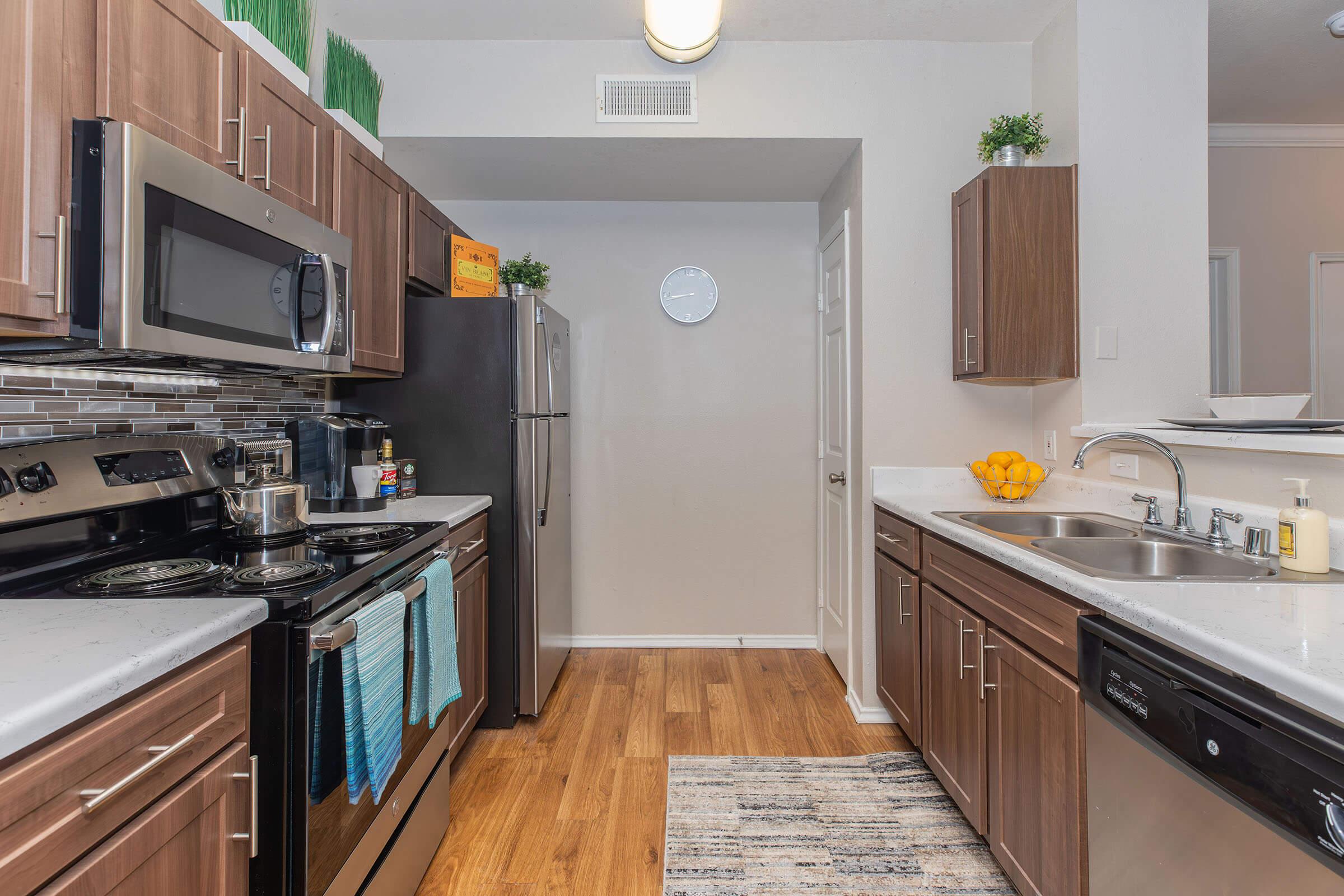 A modern kitchen featuring stainless steel appliances, including an oven, microwave, and refrigerator. The countertops are light-colored with a dark wood cabinet finish. A rug is placed on the wooden floor, and there are decorative plants and oranges on the counter for a cozy touch.