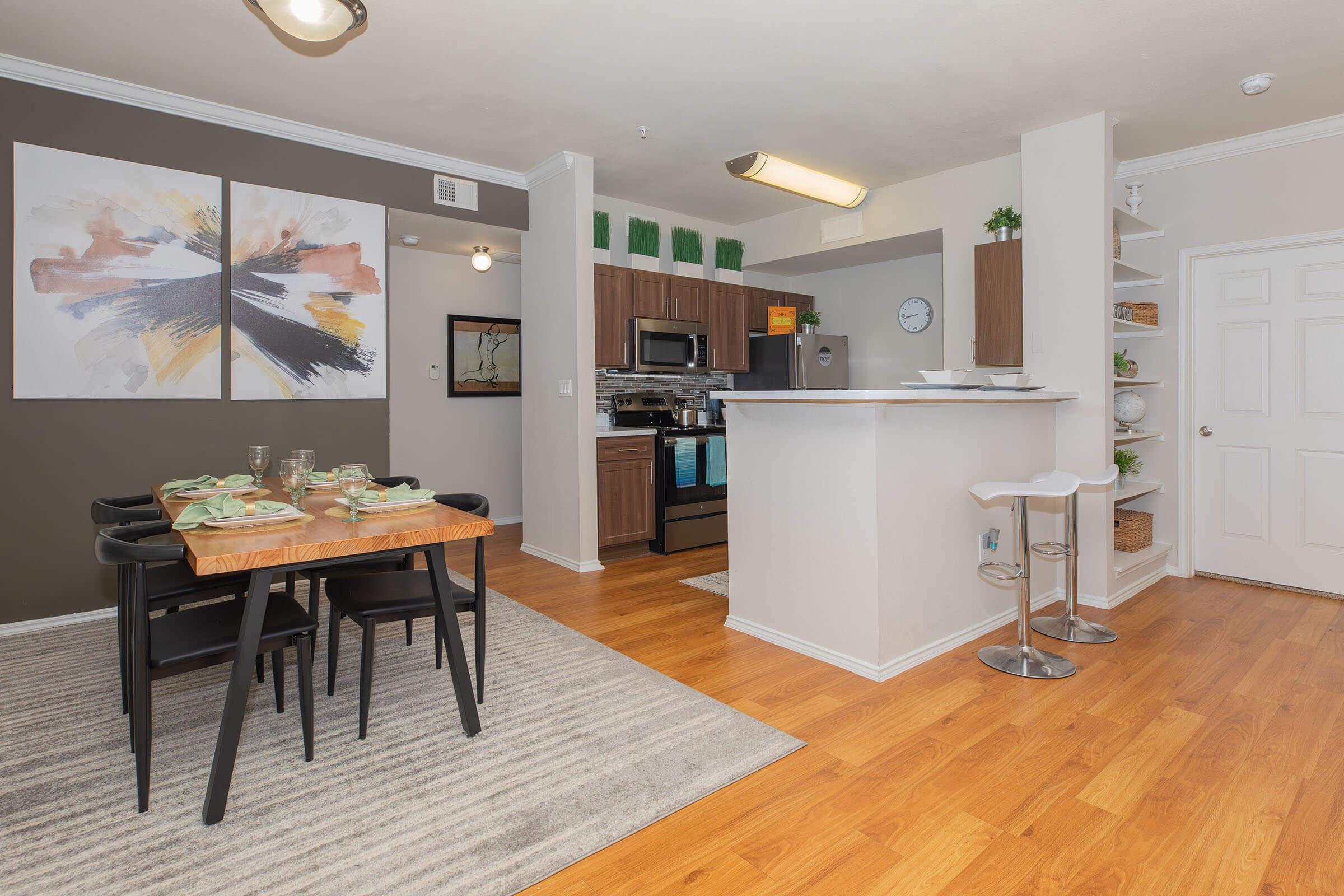 Modern kitchen and dining area showcasing a wooden dining table set for four, with green dishware. The kitchen features dark cabinetry, stainless steel appliances, and a sleek countertop. Decor includes wall art with abstract designs and a rug on hardwood flooring, creating a warm and inviting atmosphere.