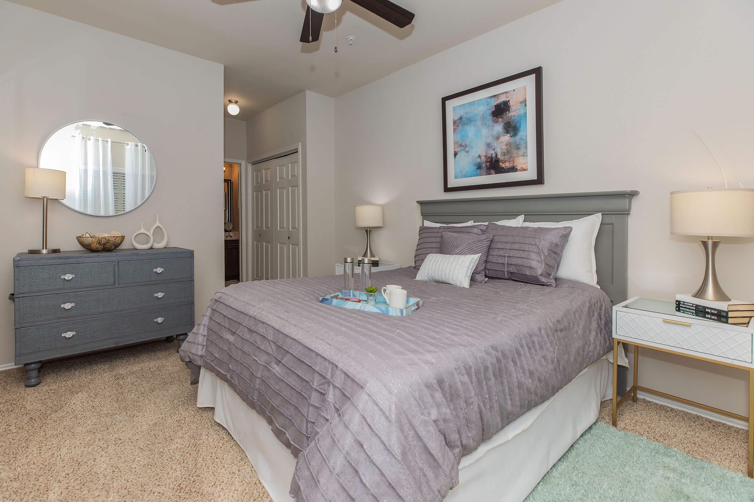A neatly arranged bedroom featuring a gray upholstered bed with purple bedding, complemented by a gray dresser and a small nightstand. A round mirror hangs above the dresser, and there's soft lighting from the lamps. The room has light-colored walls and a cozy, inviting atmosphere.