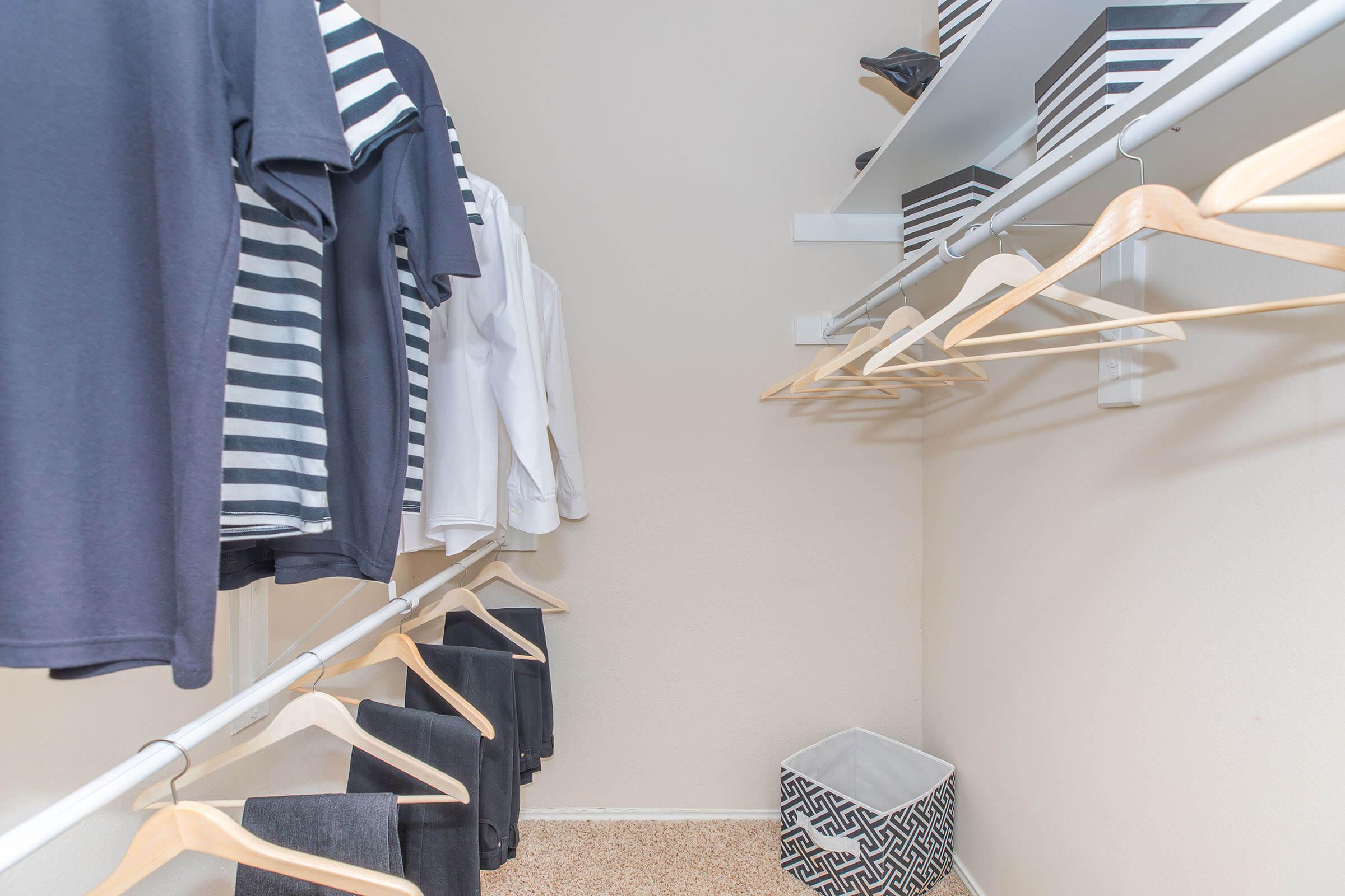 A neatly organized closet featuring various clothing items on hangers, including a striped shirt and a white blouse. The closet has a light-colored wall, a shelf with black and white storage boxes, and a decorative storage bin on the floor.