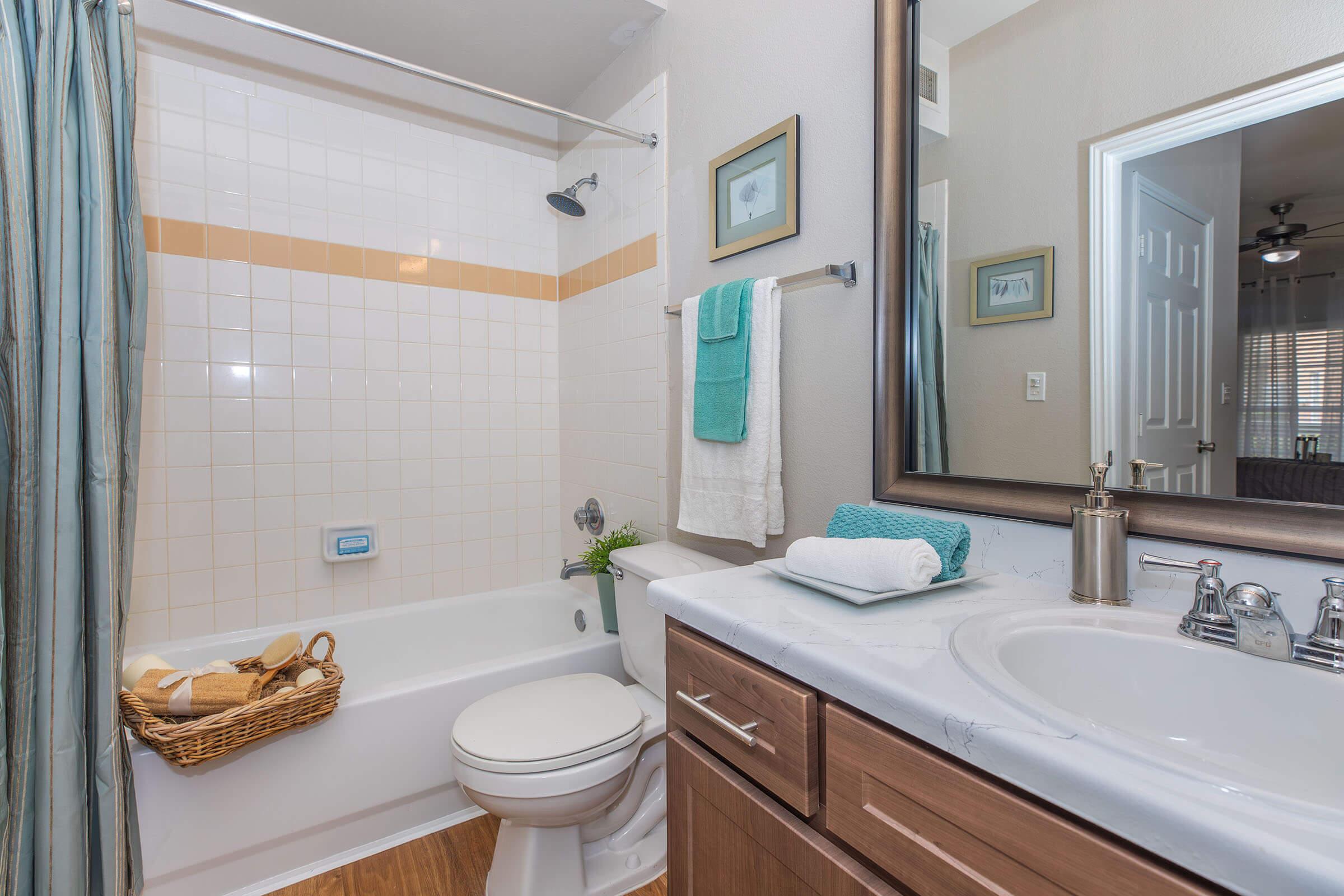 A clean bathroom featuring a white tiled shower with a light brown stripe, a bathtub with a showerhead, a toilet, and a wooden vanity with a sink. Towels are neatly hung, and a woven basket contains rolled towels. The mirror reflects the room, providing a cozy and well-organized appearance.