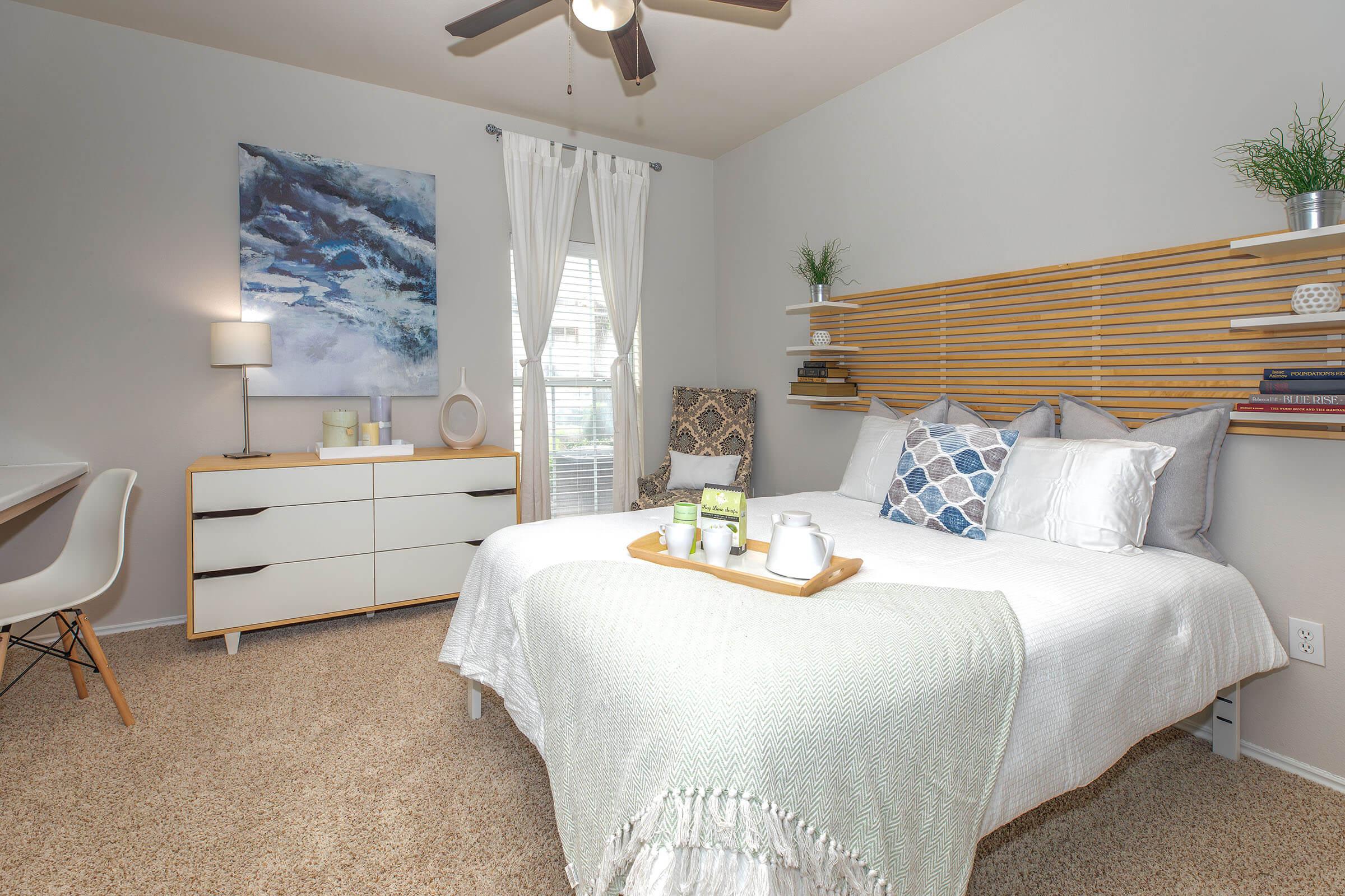 A cozy bedroom featuring a queen-size bed with decorative pillows and a soft throw. A minimalist nightstand and dresser in a light wood finish are present, along with a white desk and chair. The room has soft lighting, a fan, and wall art, creating a welcoming and stylish atmosphere.