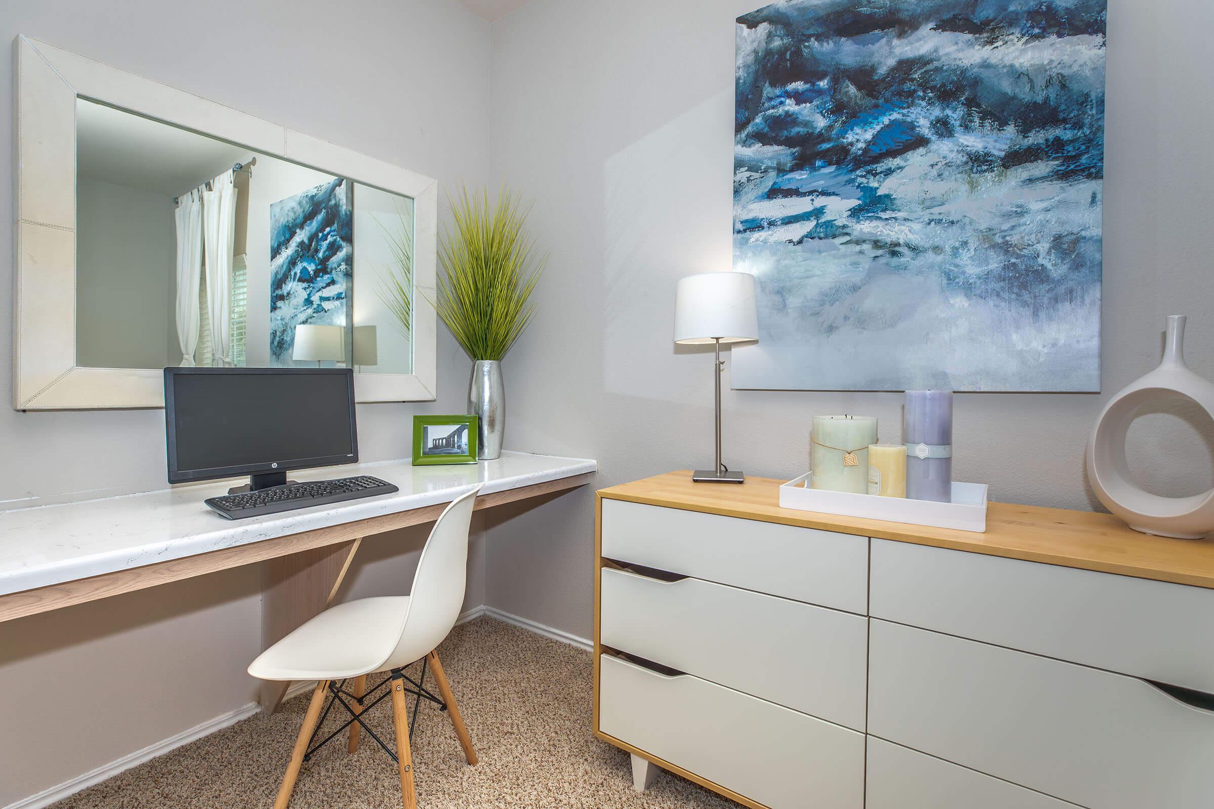 A cozy home office setup featuring a modern desk with a computer, a stylish chair, and a decorative lamp. The wall displays a large abstract painting in blue tones, and a potted plant adds a touch of greenery. A dresser with a minimalist design sits adjacent to the desk, enhancing the space's contemporary feel.