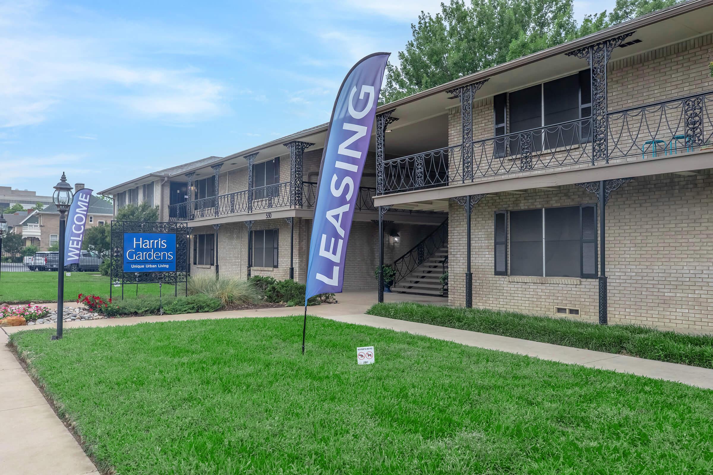 A well-maintained apartment complex with a green lawn. A "Welcome" sign and a large "Leasing" banner are prominently displayed. The building features two stories with outdoor staircases, and there are colorful flower beds at the entrance.