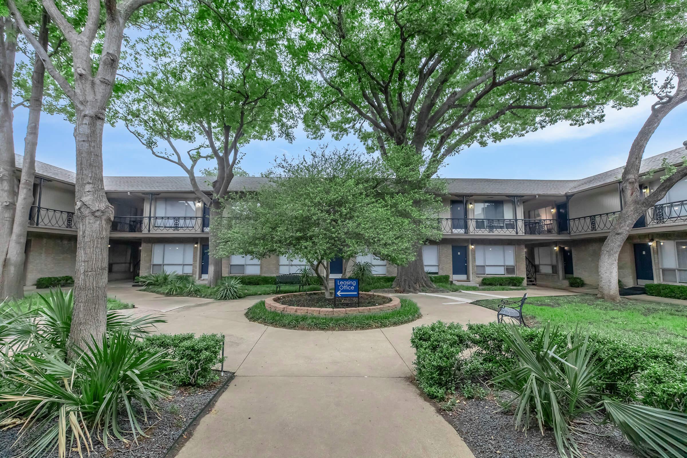 A courtyard view of an apartment complex featuring lush green trees, well-maintained landscaping with shrubs and grass, and two-story buildings with balconies. A sign indicating "Garden Apartments" is visible. The scene is peaceful with a clear sky, creating a welcoming atmosphere.