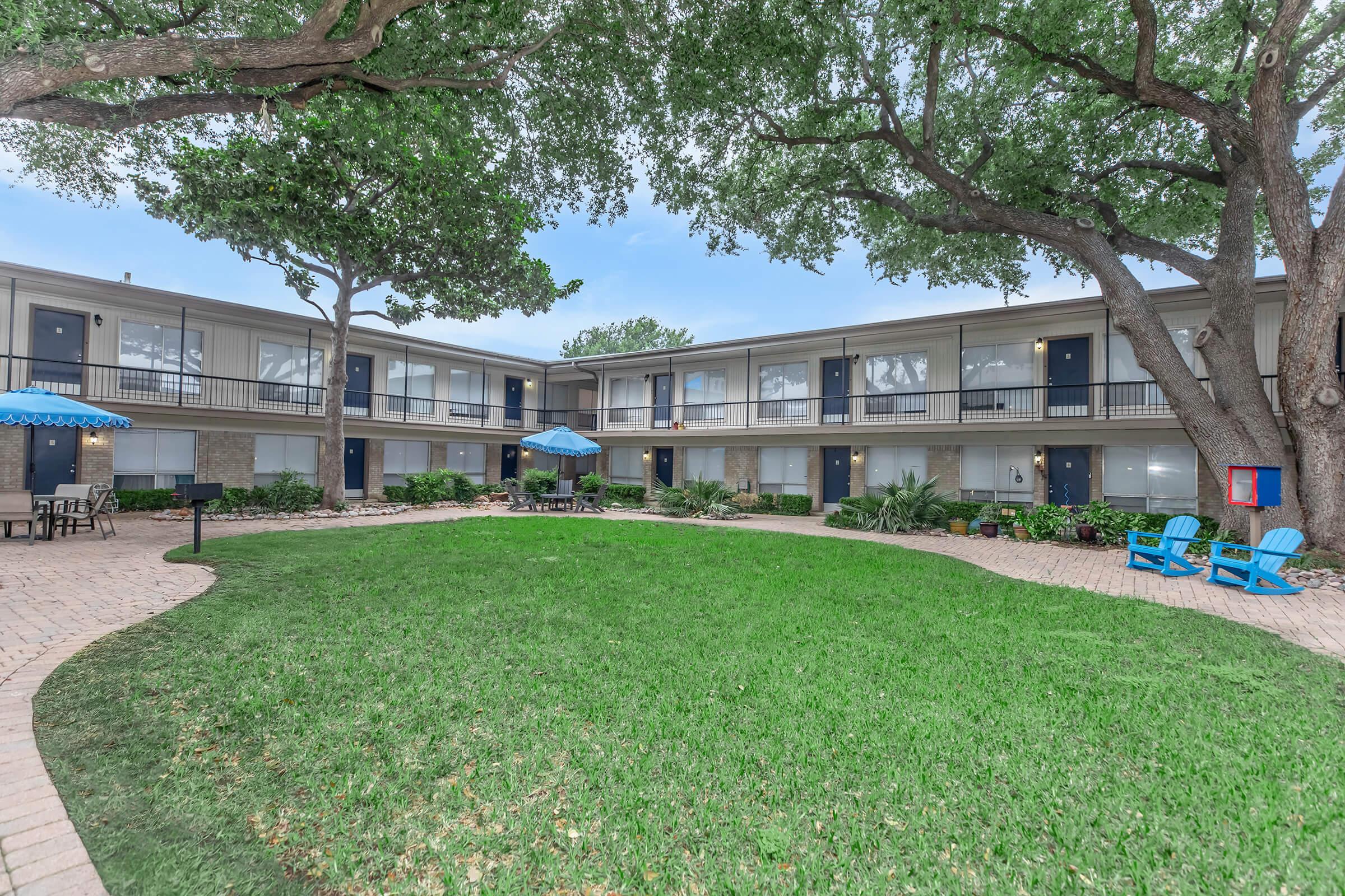 A view of a courtyard at a multi-unit building, featuring green grass, several trees, and outdoor seating with blue chairs. In the center, there is a small patio area with an umbrella and tables. The building has multiple levels with balconies and is surrounded by landscaping.