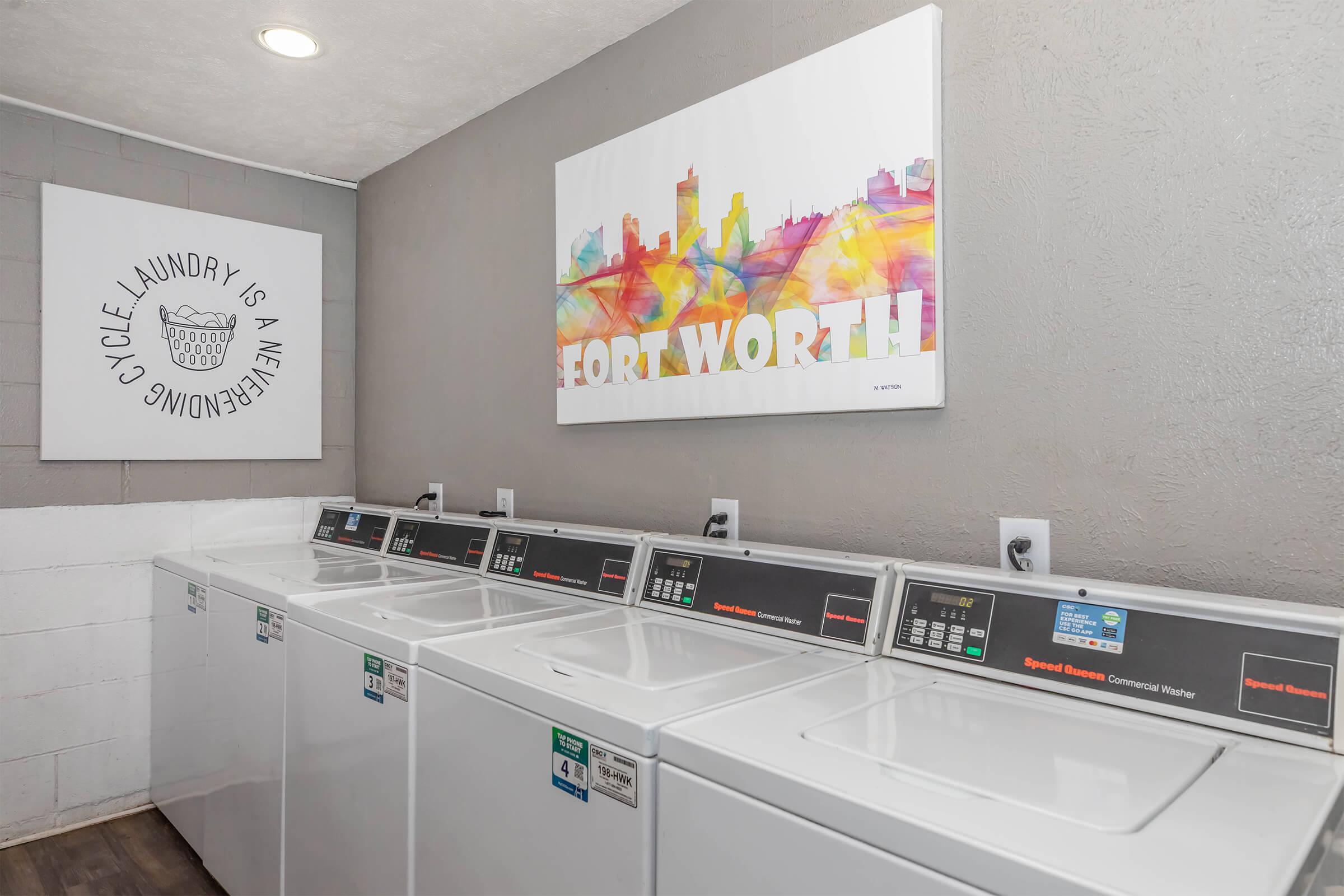 A modern laundry facility featuring several commercial washing machines lined up against a wall. To the left, there's a decorative wall art piece with the text "Laundry is an Adventure" and on the right, a colorful artwork depicting the skyline of Fort Worth. The walls are painted in neutral tones.