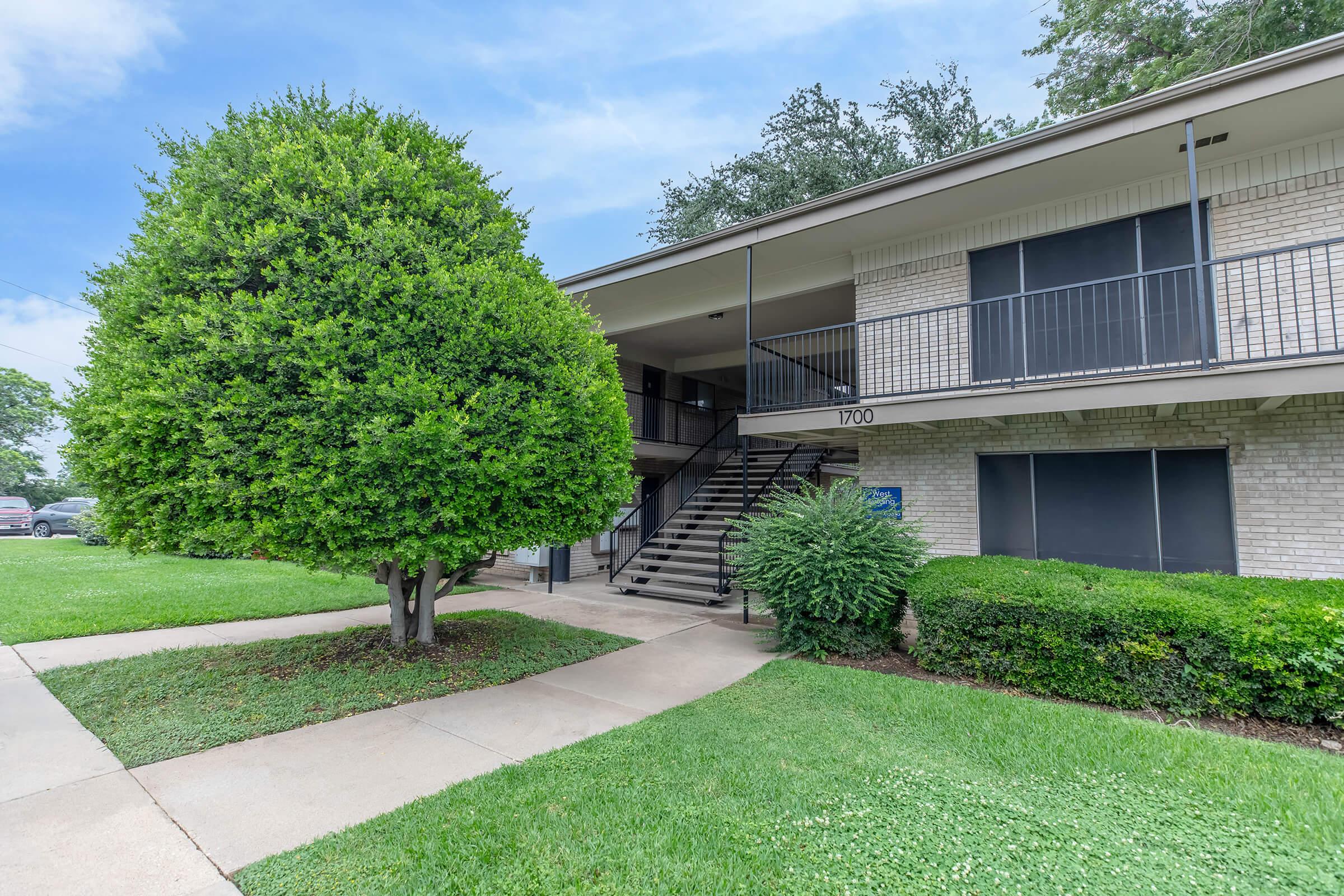 A two-story apartment building with a staircase leading to the second floor. The exterior features brick walls, a well-maintained lawn with a large bushy tree and hedges. The building number is 1700, and the sky is partially cloudy.