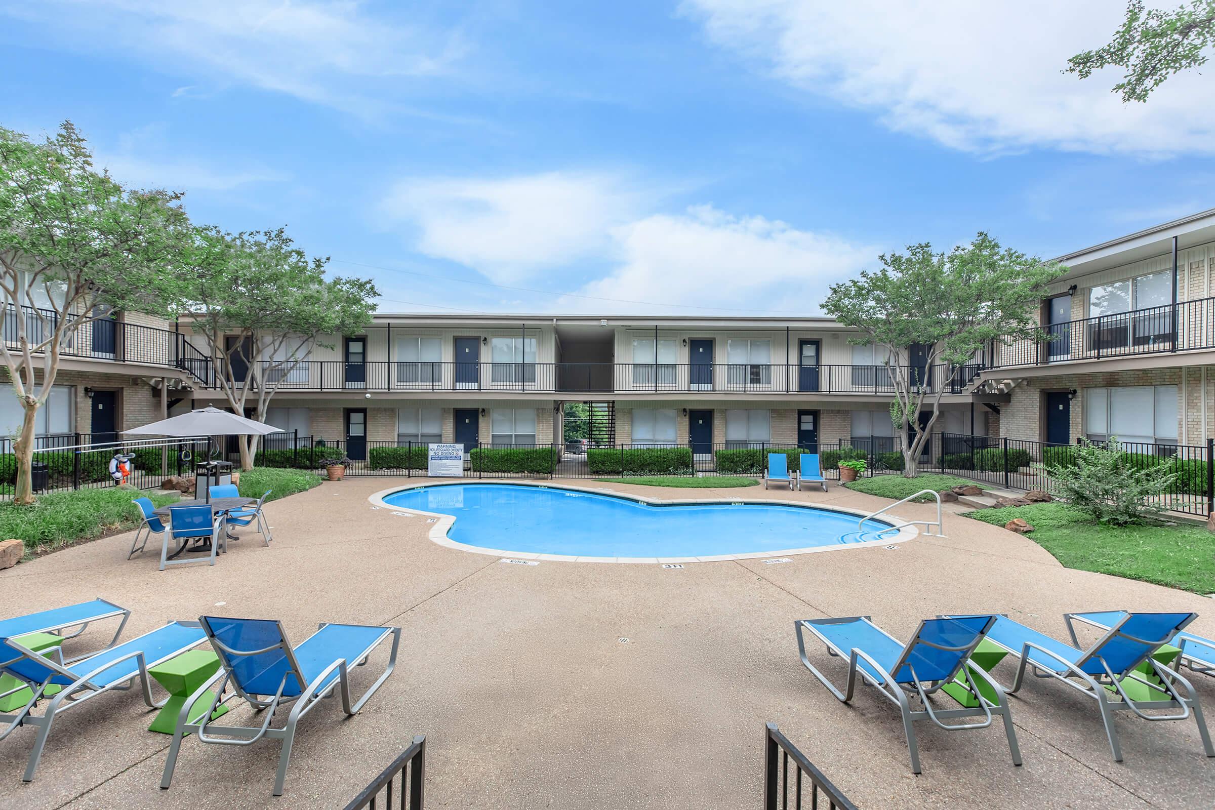 A tranquil courtyard featuring a swimming pool surrounded by lounge chairs. Green shrubs and trees provide shade, while adjacent to the pool, there are umbrellas for relaxation. Two-story building units encircle the pool area, creating a serene atmosphere. Bright, clear sky overhead adds to the inviting scene.