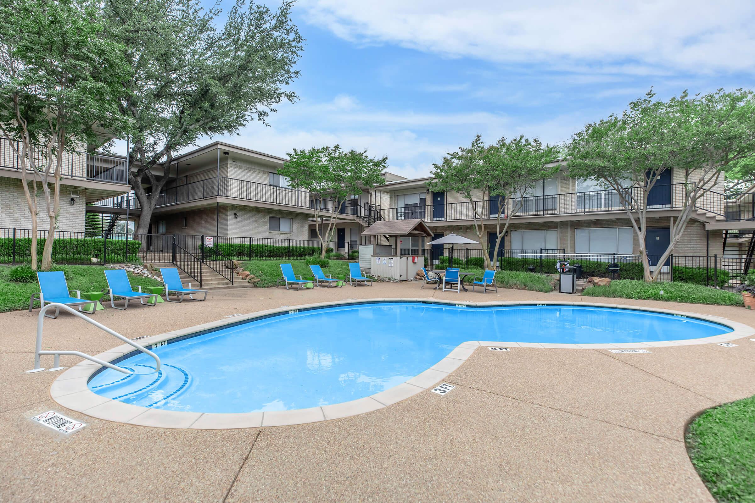 A well-maintained outdoor swimming pool surrounded by lounge chairs. The area features greenery with trees and two-story apartment buildings in the background. A shaded gazebo is visible near the poolside, creating a relaxing atmosphere. The sky is partly cloudy, enhancing the inviting setting.