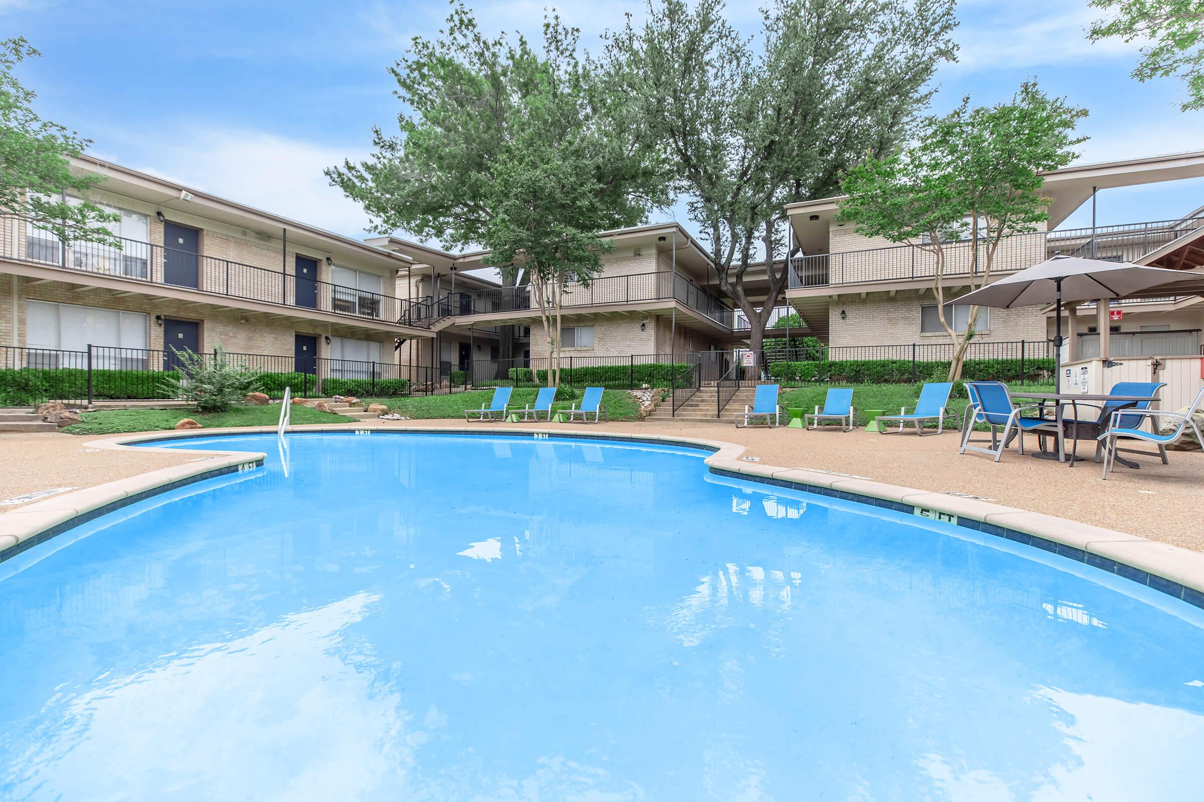 A clear blue swimming pool surrounded by lounge chairs and umbrellas, with a landscaped area featuring trees and shrubs. In the background, there are two-story apartment buildings with balconies. The scene is bright and inviting, perfect for relaxation and leisure.