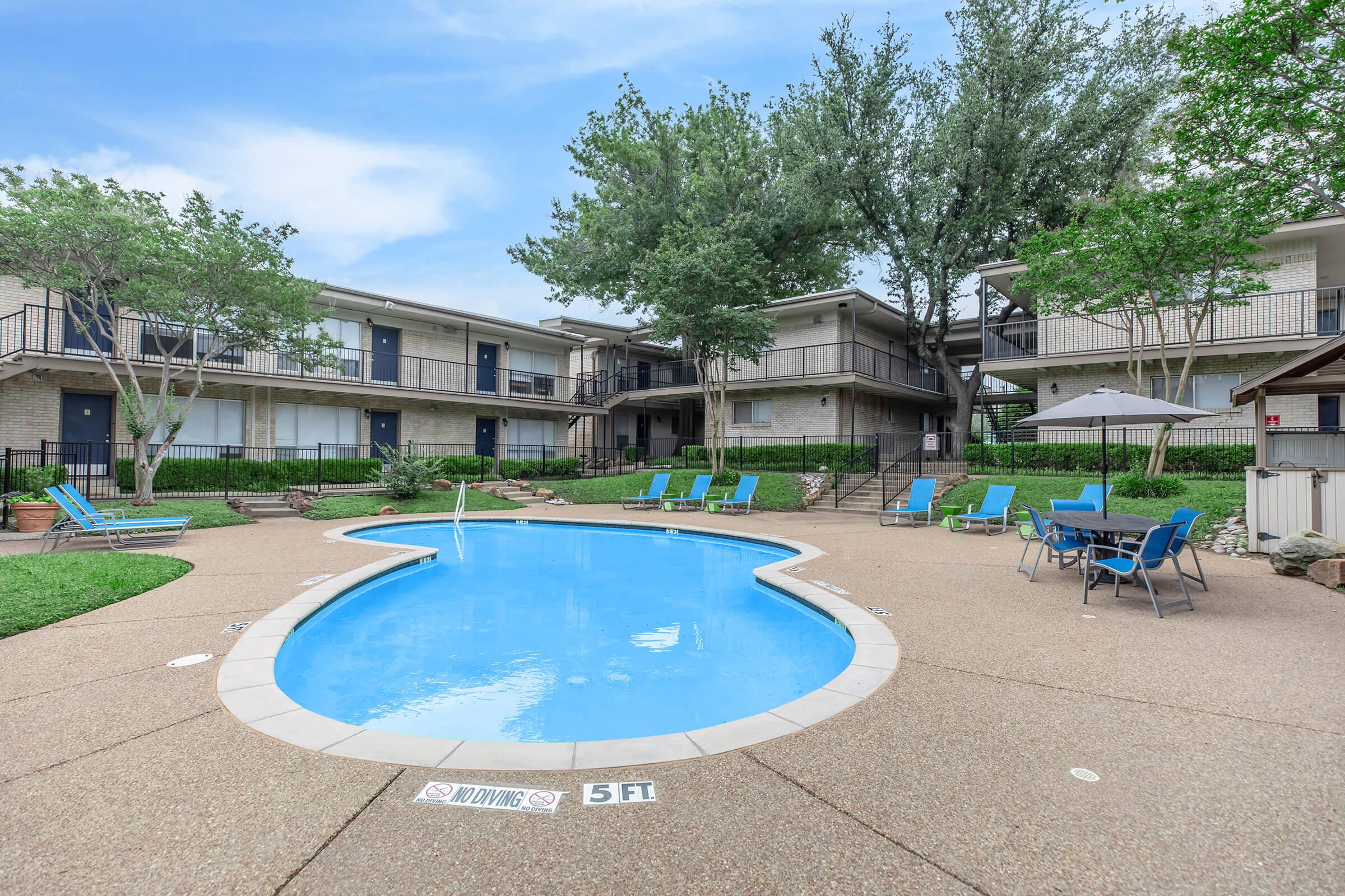 A swimming pool in a courtyard surrounded by residential buildings. The pool features lounge chairs nearby and is marked with a depth sign. Lush greenery is visible around the area, with several trees providing shade. Umbrellas are set up for additional seating comfort.