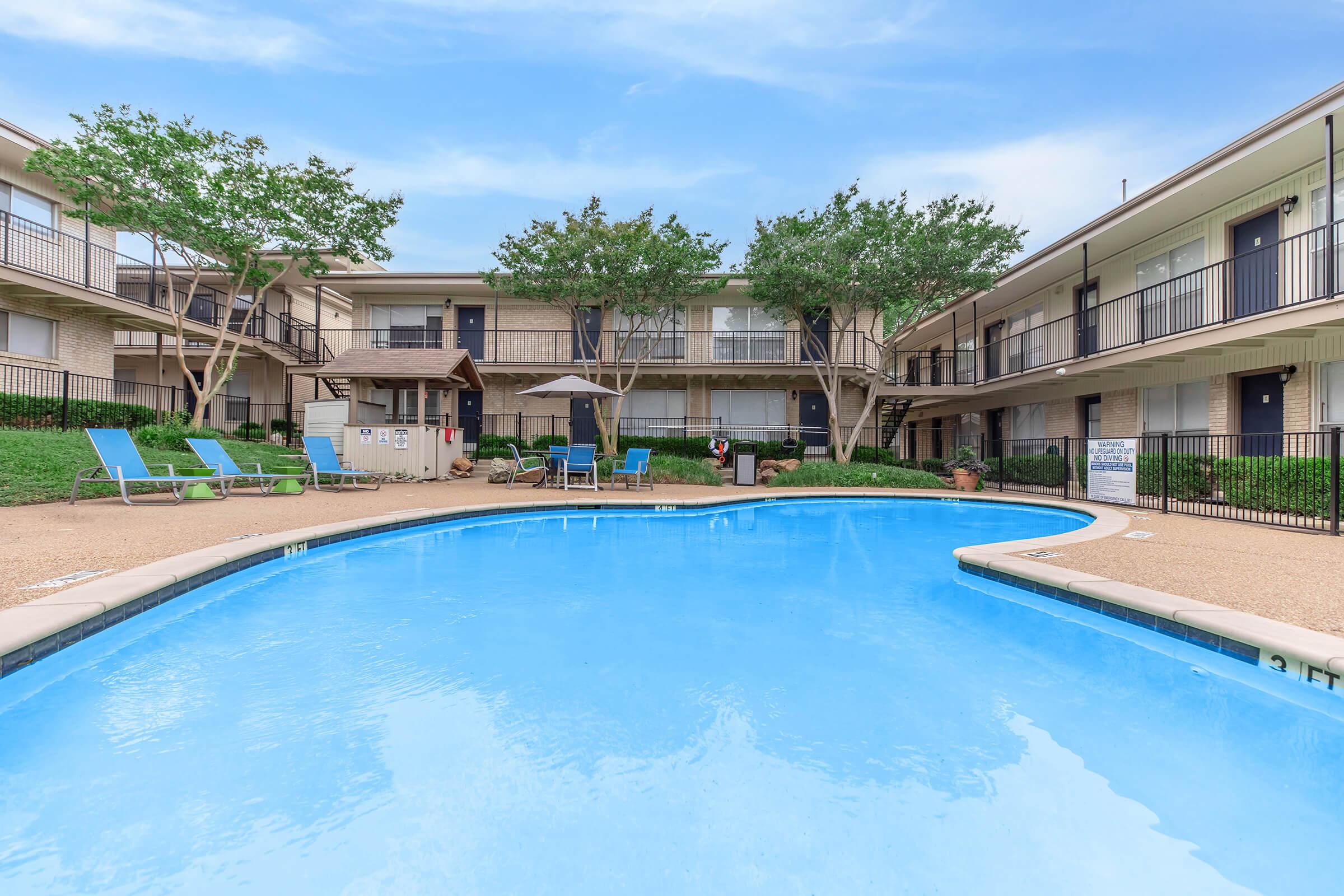 A clear blue swimming pool surrounded by lounge chairs and shade structures, located in a courtyard area flanked by two-story apartment buildings with balconies. Lush green trees are visible in the background under a bright blue sky.