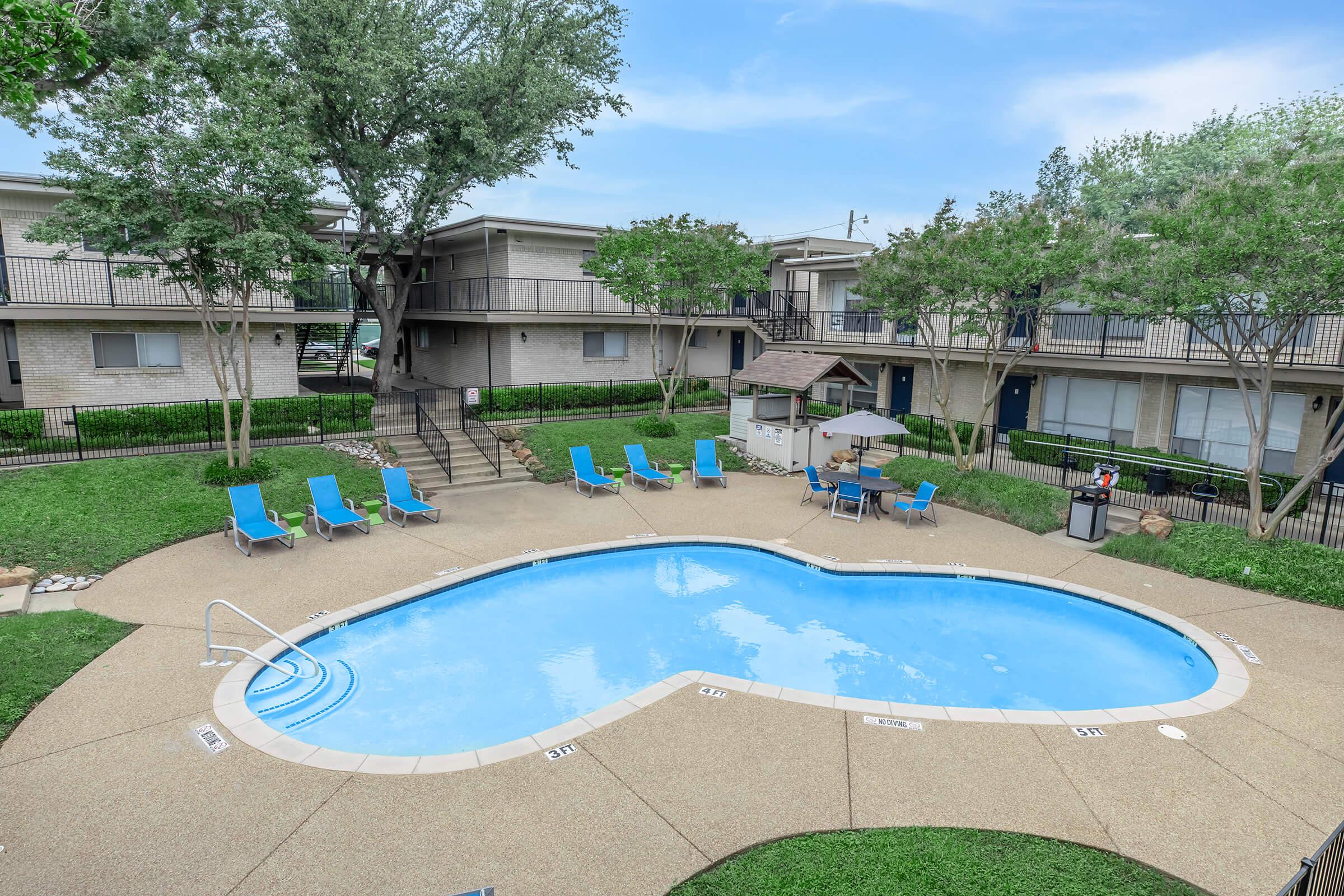 A heart-shaped swimming pool surrounded by green grass and trees. Lounge chairs are arranged around the pool, and a shaded seating area is visible. Nearby buildings feature balconies and a clear blue sky above.