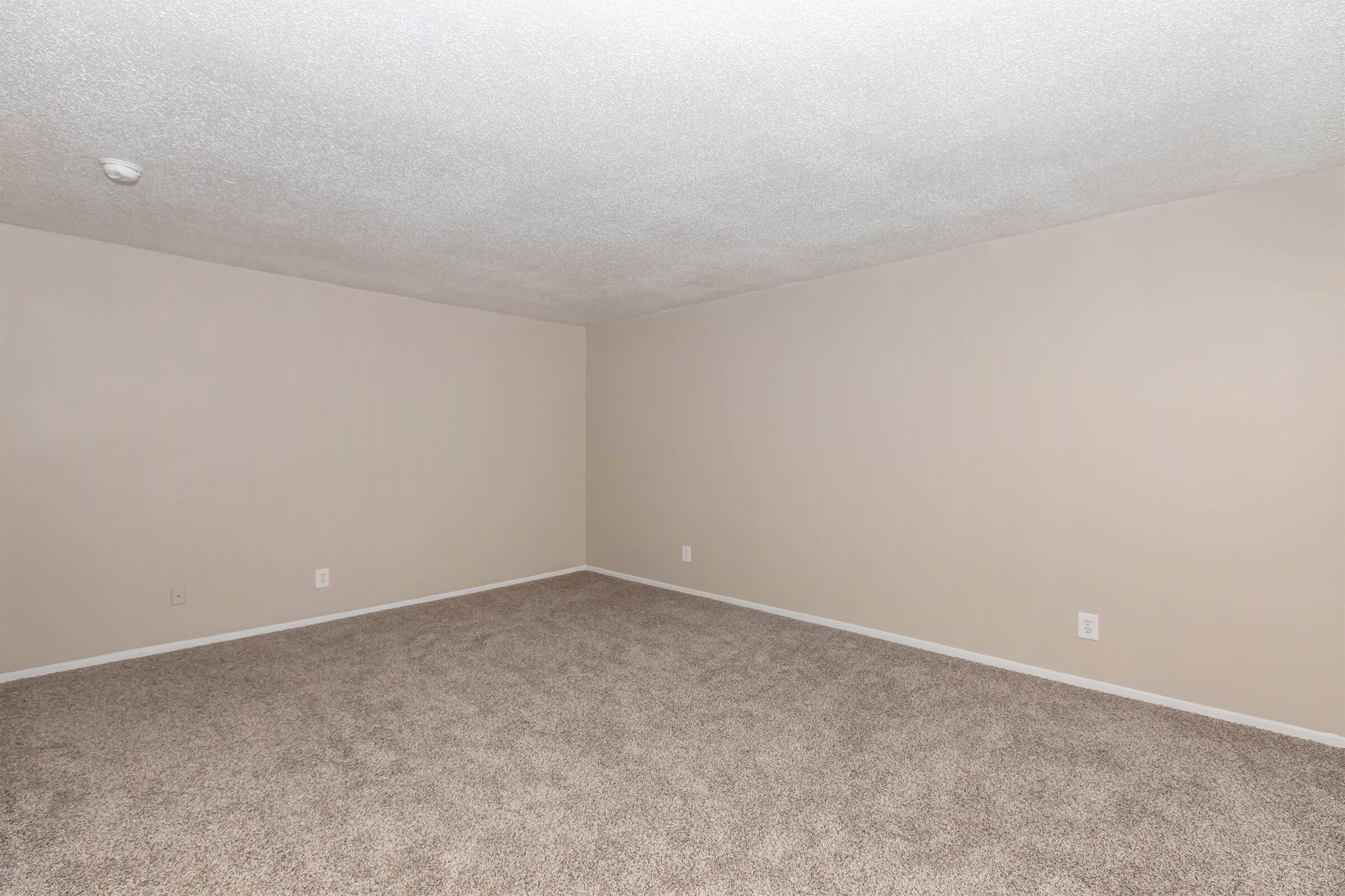 Empty room with beige walls and a light carpet. The ceiling is textured, and there are no furnishings or decorations. The space is well-lit, creating a clean and open atmosphere.